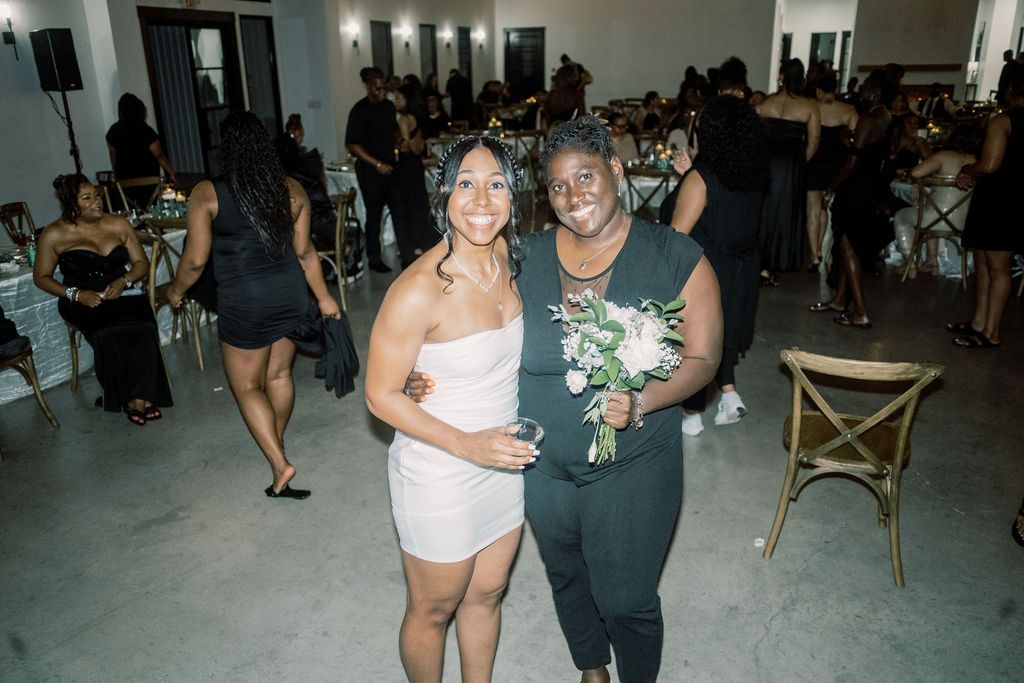 Two women posing together at an elegant wedding reception, one holding a bouquet, surrounded by guests in formal attire.