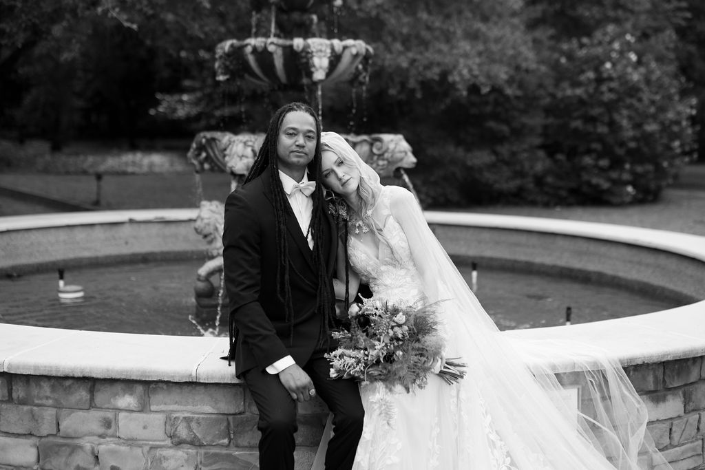 Black and white wedding photo of bride and groom posing by a stone fountain in a garden setting.