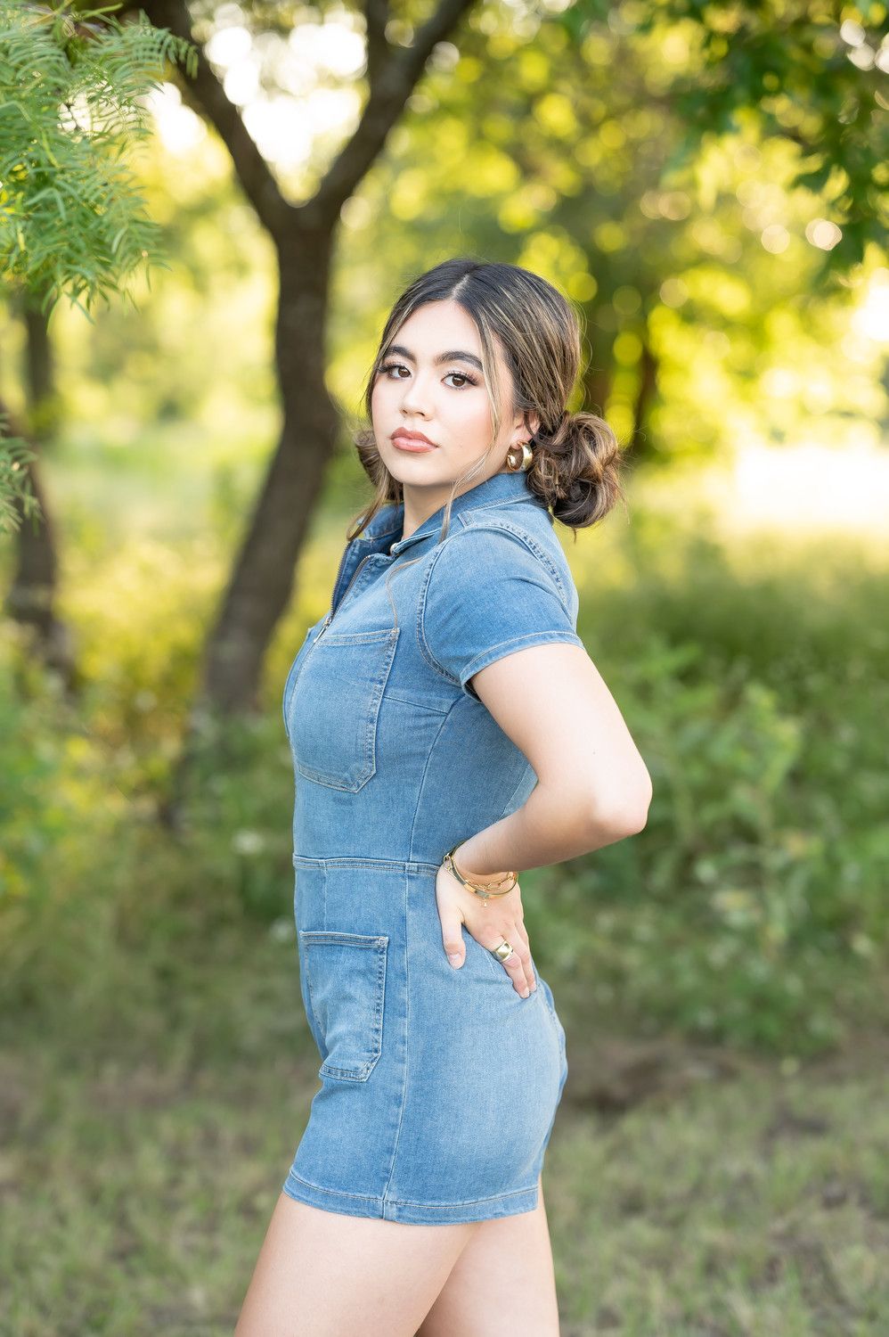 Young woman in denim romper posing outdoors among green trees in a sunlit park. Wichita Falls & the surrounding North Texas