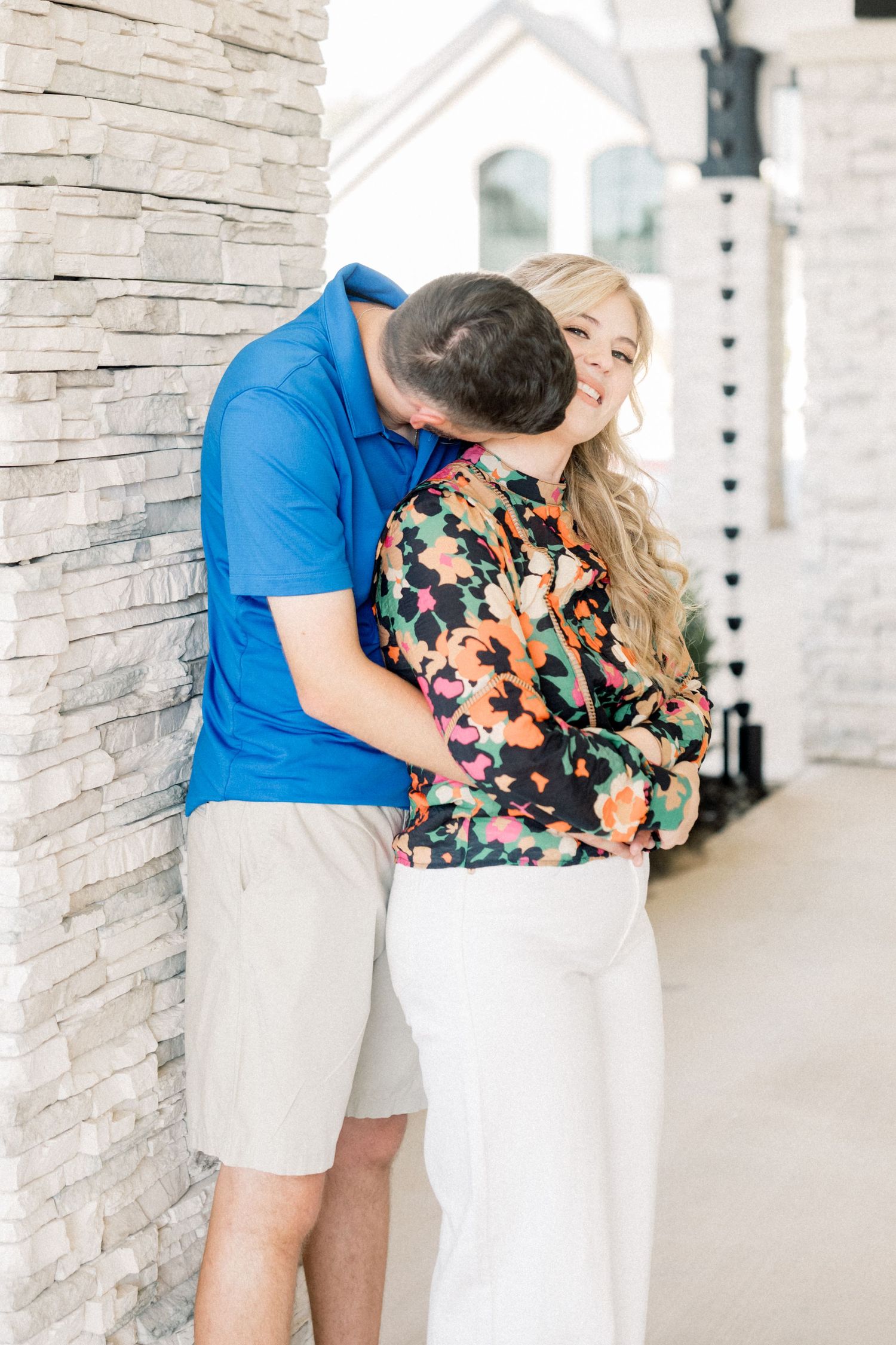 Man in blue polo shirt kissing woman in floral top against a stone wall exterior.