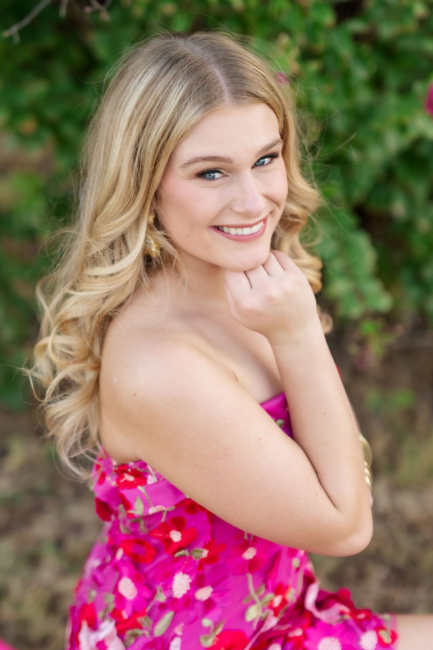 Young blonde woman in pink floral strapless dress smiling outdoors with green foliage background.