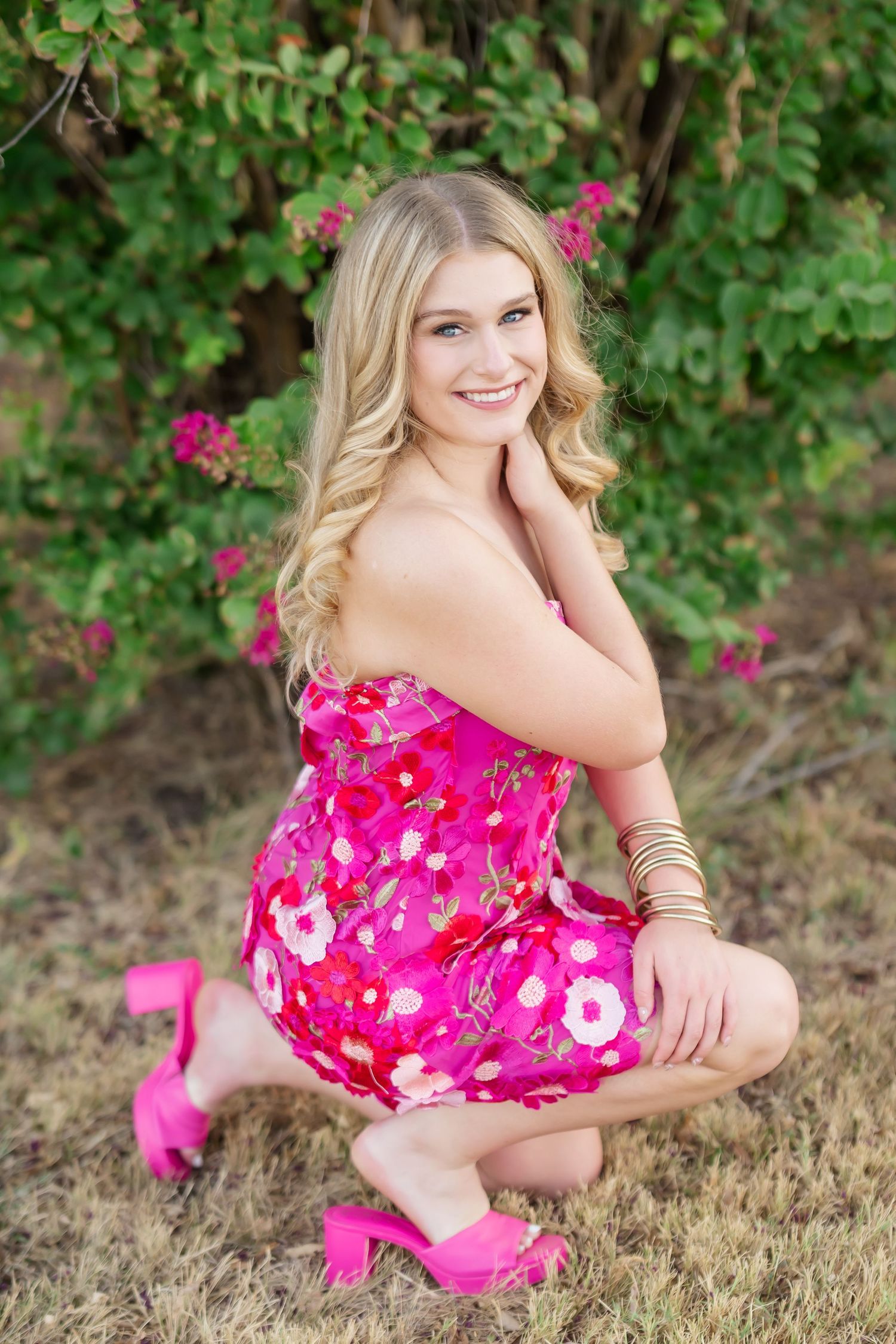 Smiling blonde teen girl in pink floral strapless dress and heels crouching near flowering bushes outdoors.