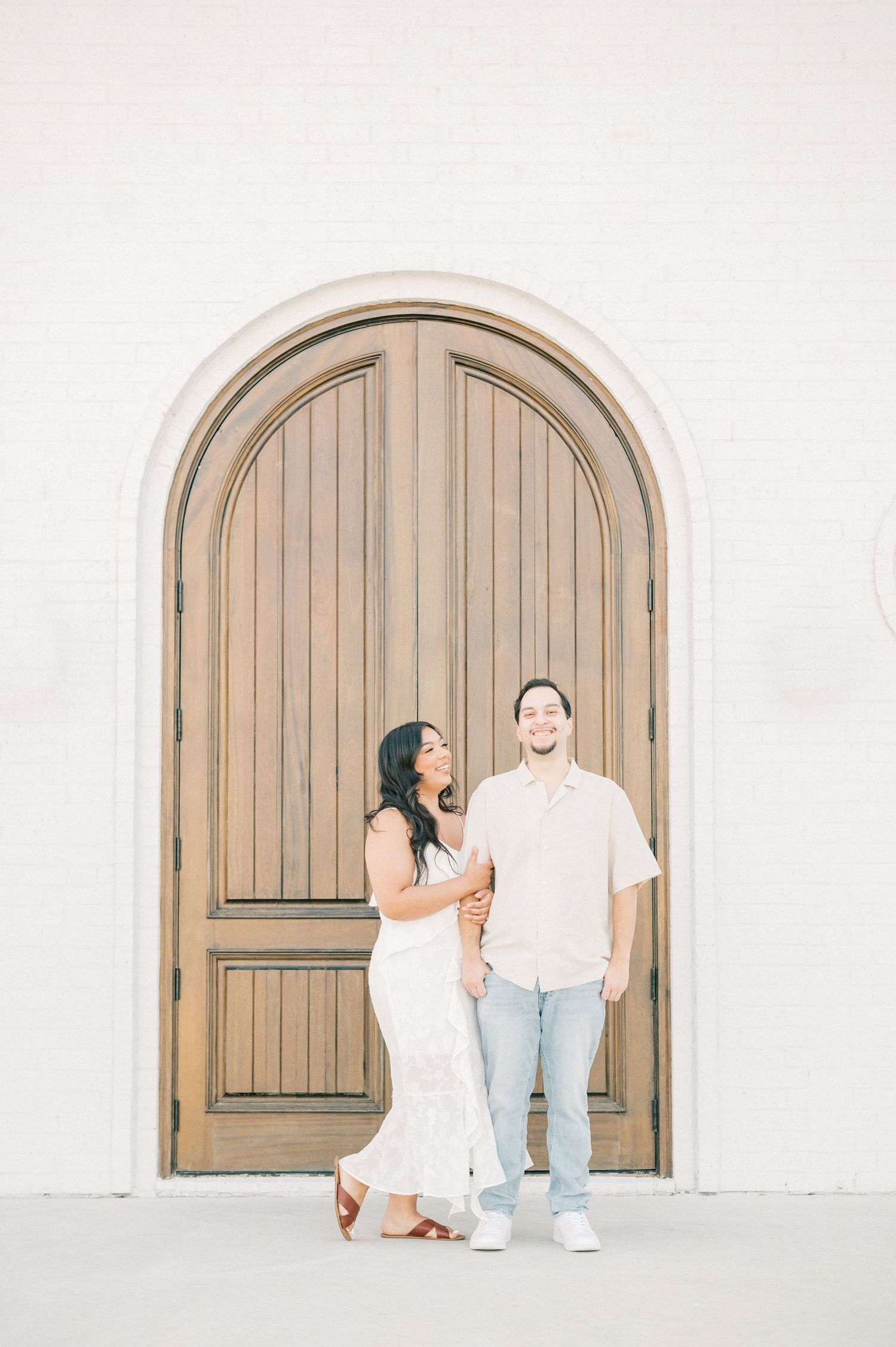A couple stands in front of an arched wooden door against a white wall during an engagement photo session.