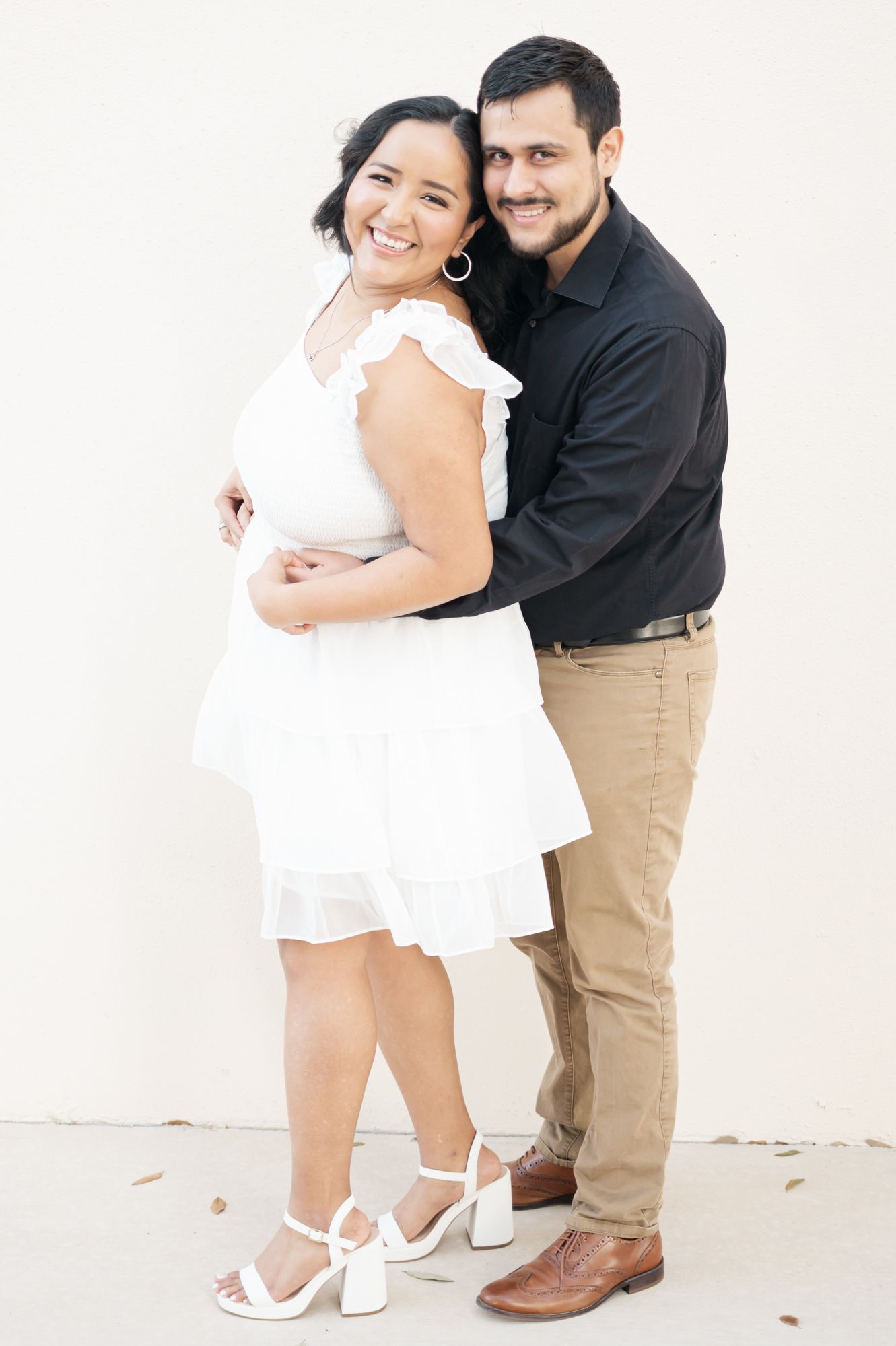 A couple poses together against a white wall, with the woman wearing a white dress and the man in a black shirt.