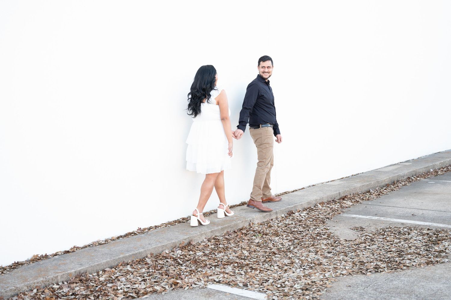 A couple takes creative engagement photos against a minimalist white wall backdrop.