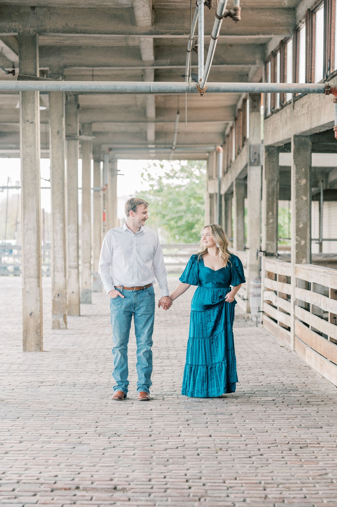 A couple walks hand in hand through an industrial covered walkway, with the woman wearing a flowing teal dress.