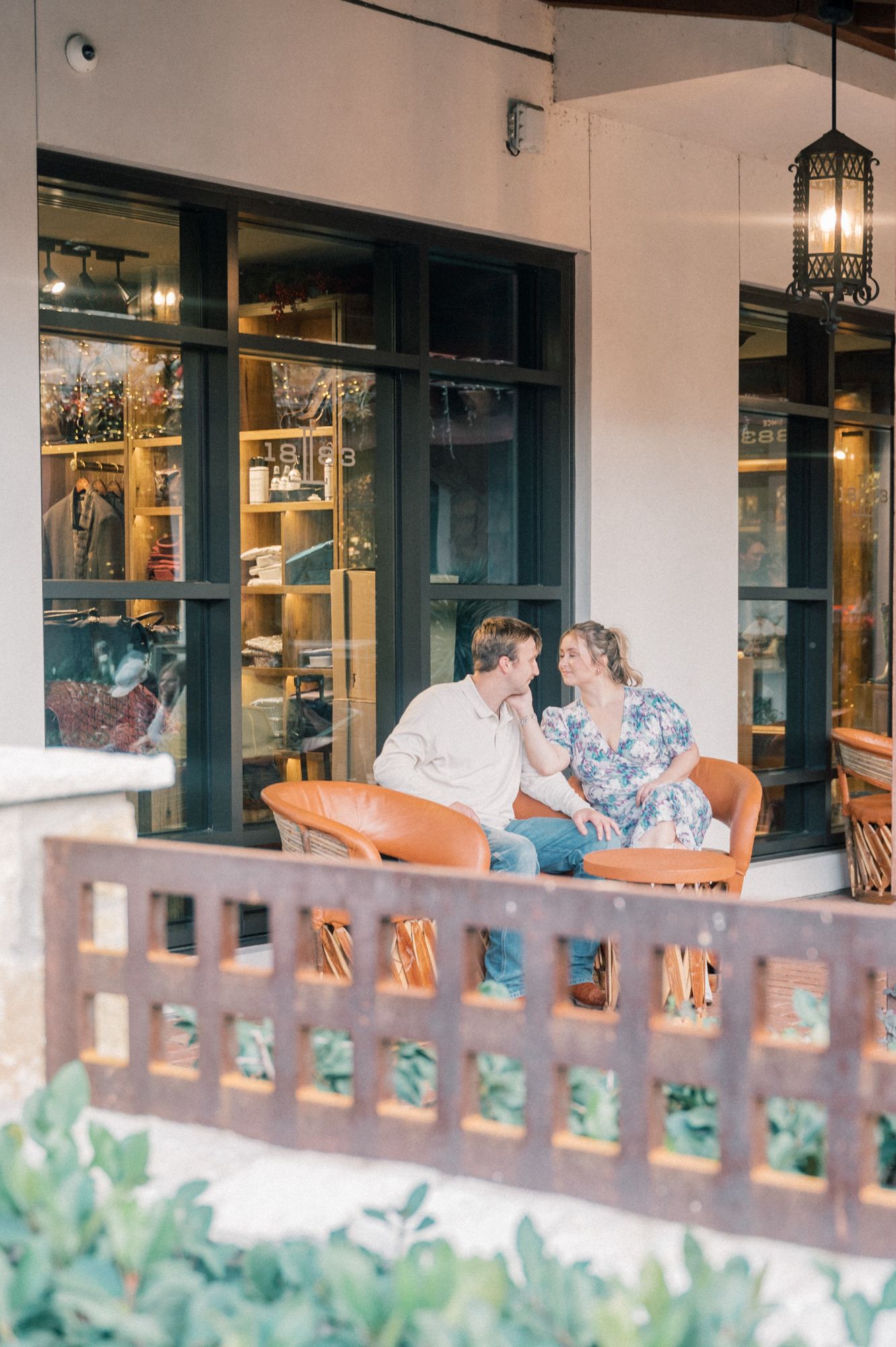 Couple sitting on rustic wooden chairs on a covered patio with brick flooring and decorative lighting.