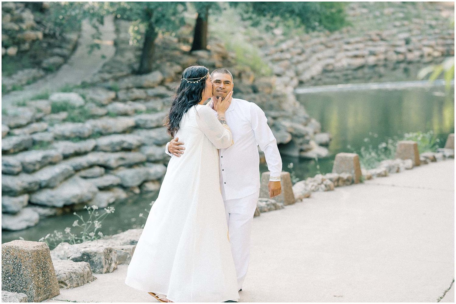 A couple in white clothing embraces on stone steps surrounded by natural scenery.