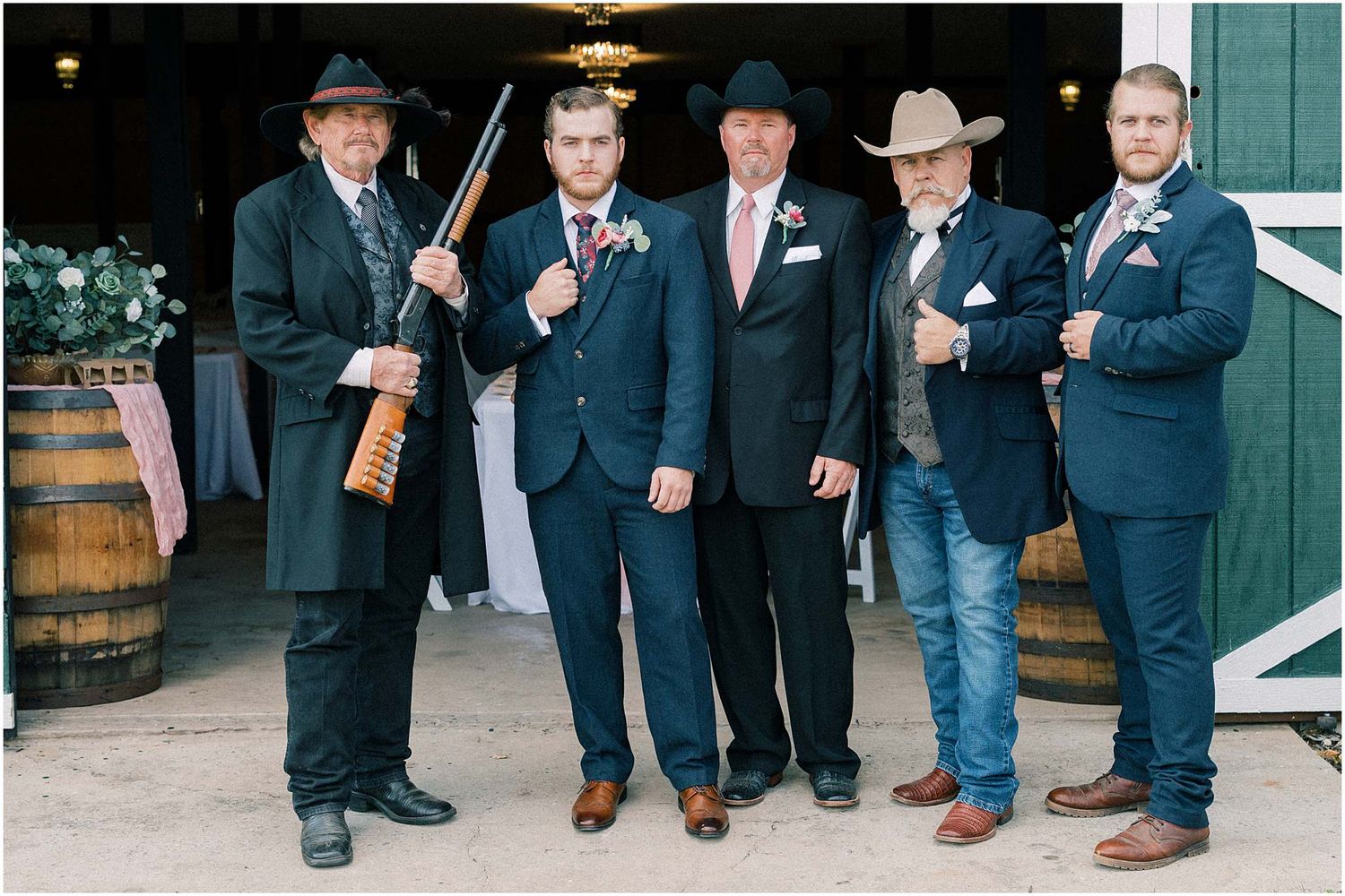 Groomsmen in dark suits pose together at rustic venue entrance.