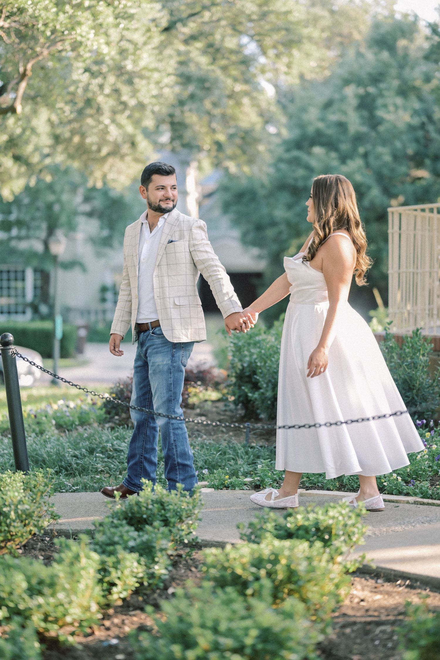 Couple holding hands while walking along a garden path, the woman in a flowing white dress leading the man in jeans and blazer.