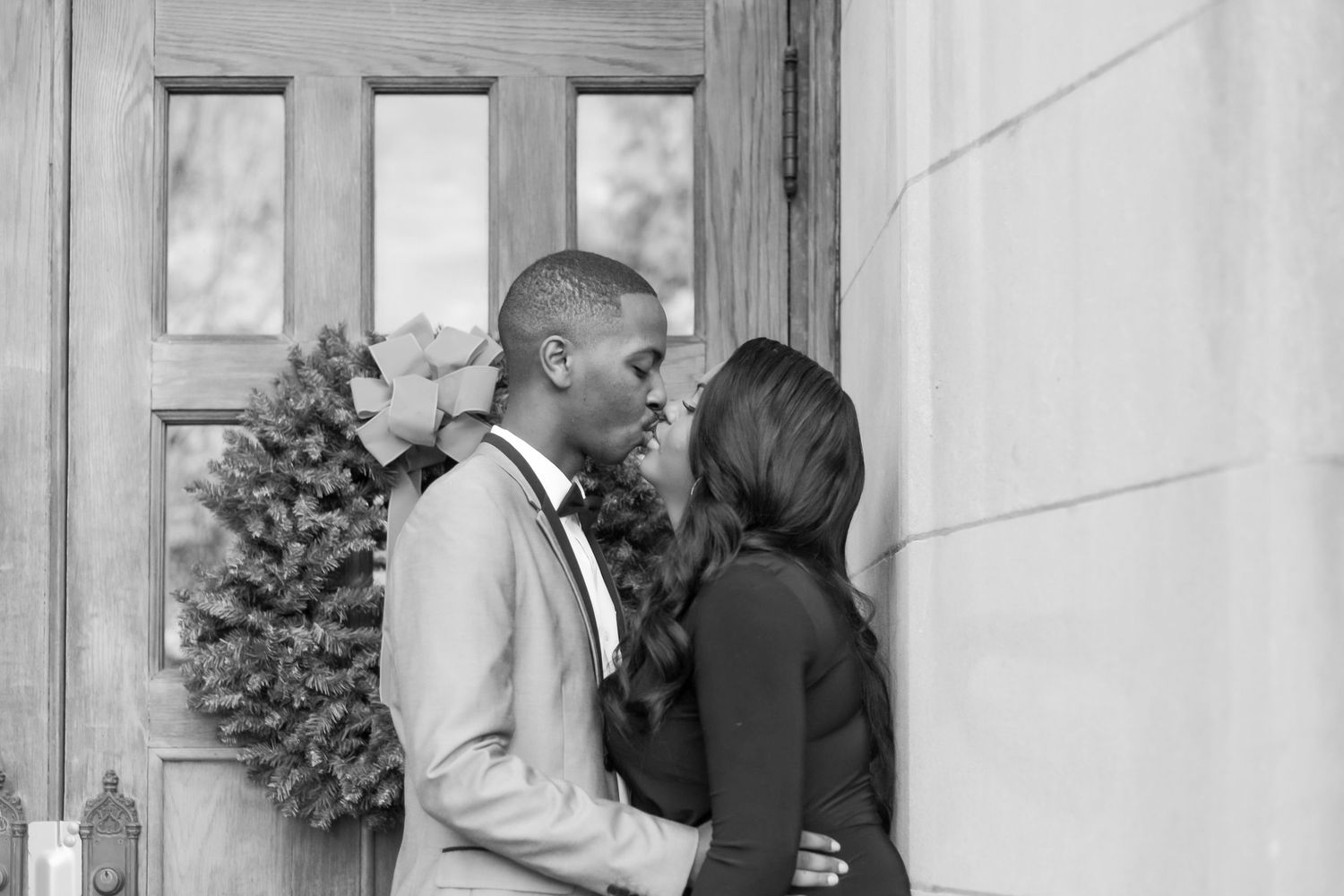Black and white photo of a couple sharing a romantic moment by a wooden door with a Christmas wreath.