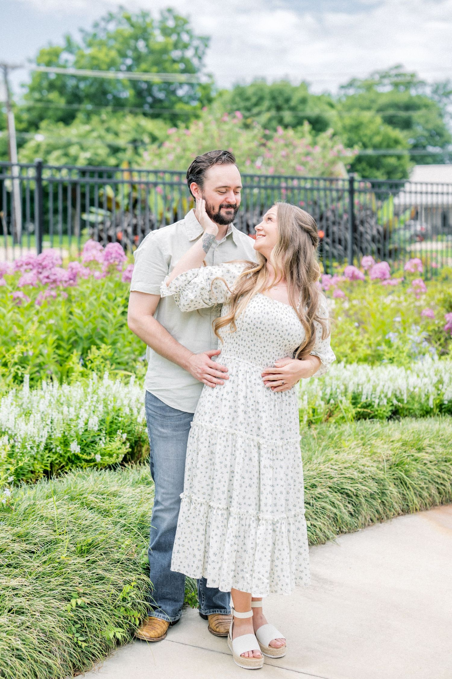 Couple embracing and smiling in a colorful garden, woman in white floral dress and man in gray shirt.