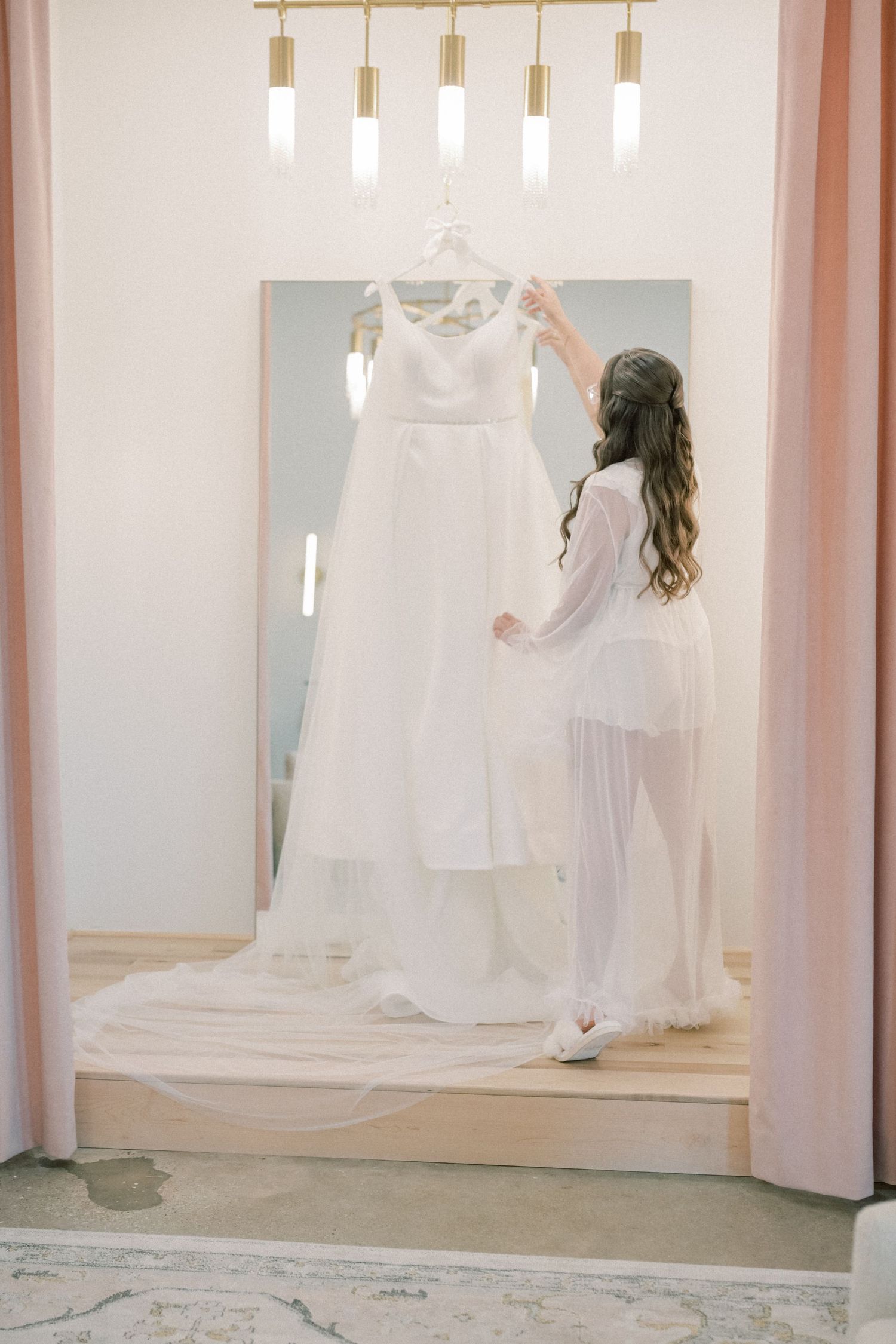 Bride in white robe admires her wedding gown hanging in a softly lit bridal suite.