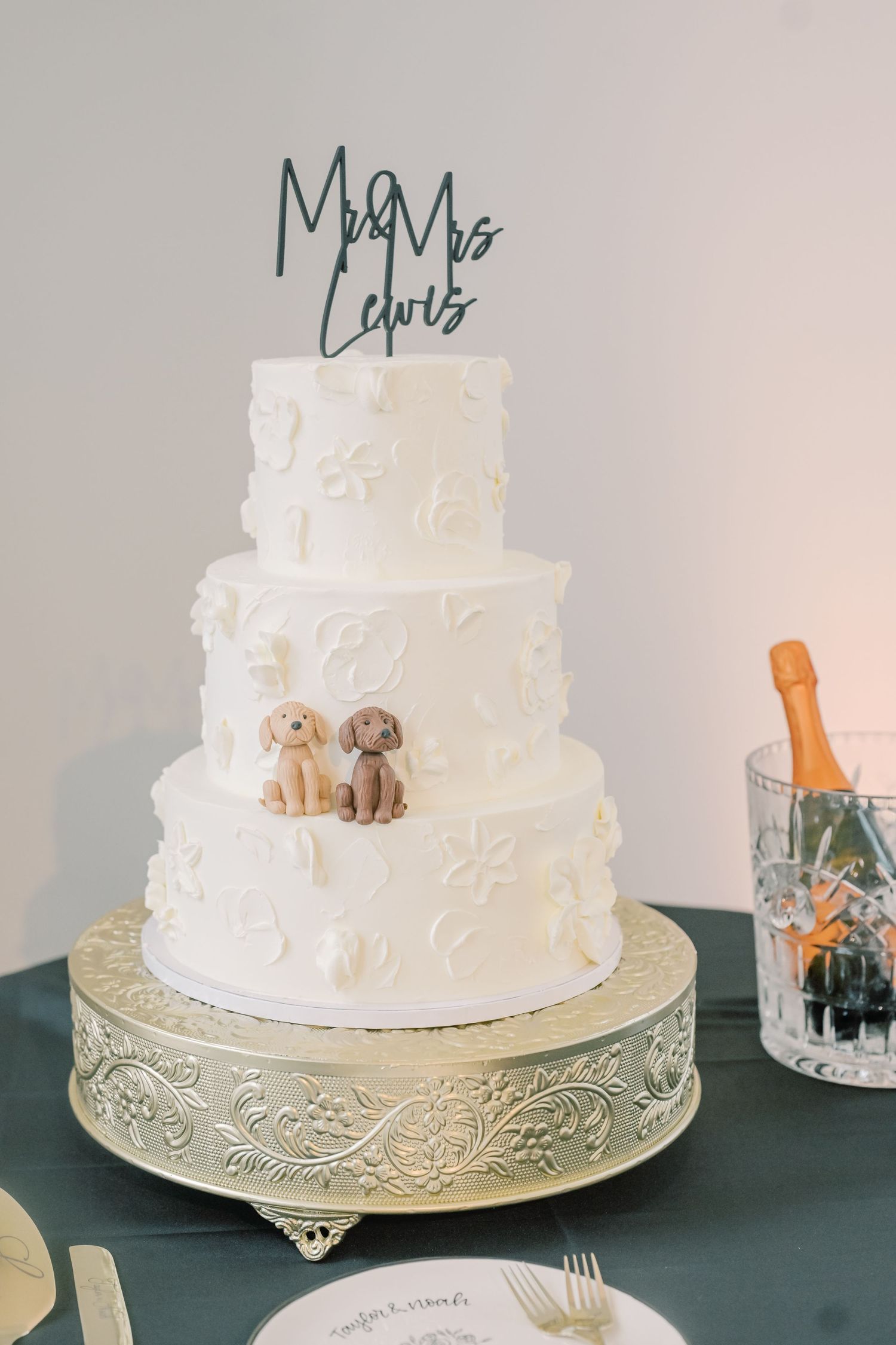 Elegant three-tier white wedding cake with puppy figurines and Mr & Mrs Lewis topper on gold stand.