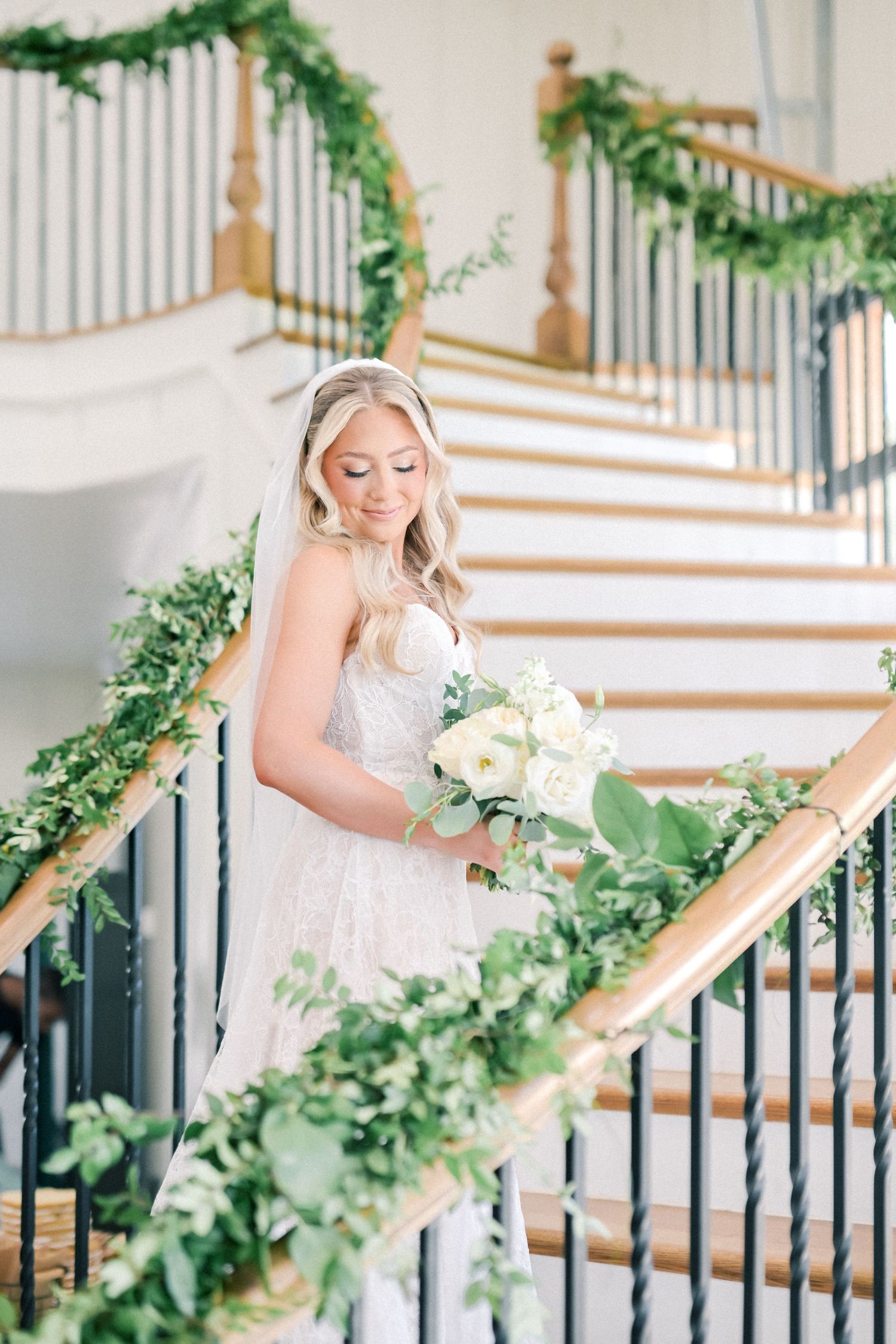 Bride in lace gown holding white bouquet on staircase decorated with green garland.