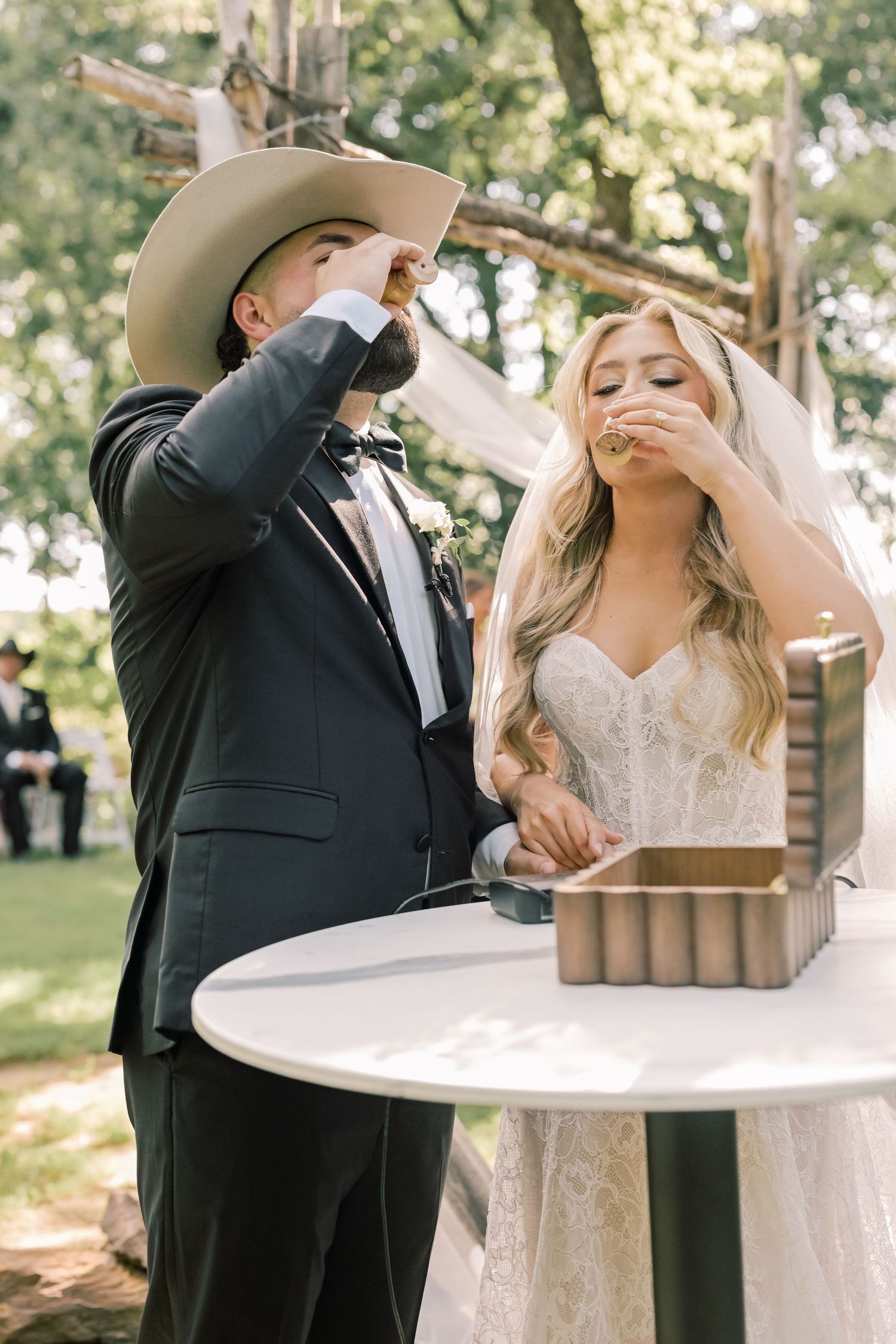 Bride and groom in cowboy hat performing unity ceremony drinking shot at outdoor wedding.