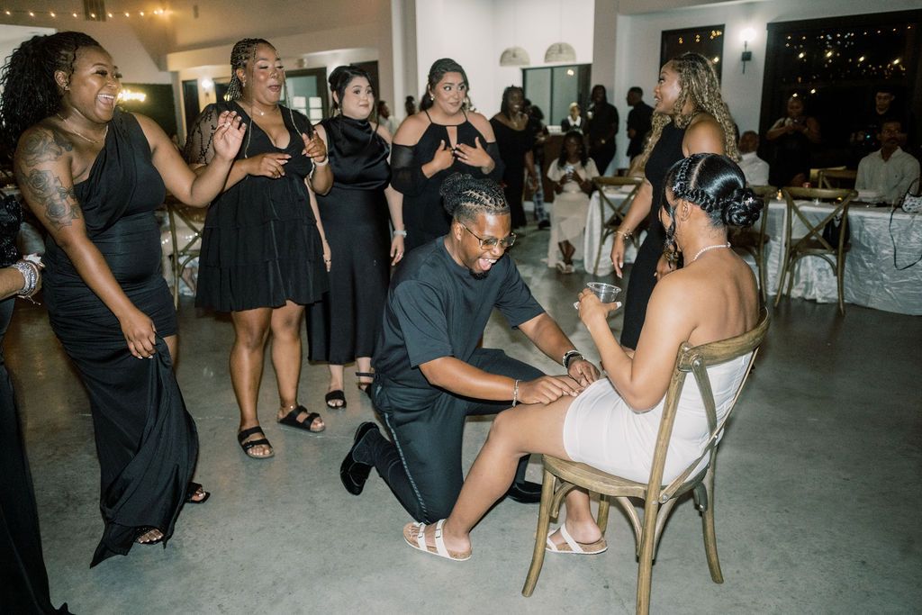 Groom kneeling before bride in white dress, surrounded by bridesmaids in black at a wedding reception.