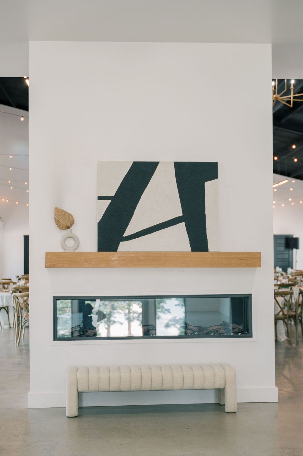 Modern white fireplace with wood mantel, abstract art, and tufted bench in elegant event venue.