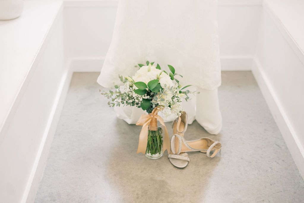 White bridal bouquet with greenery in glass vase beside nude strappy heels against flowing wedding dress.