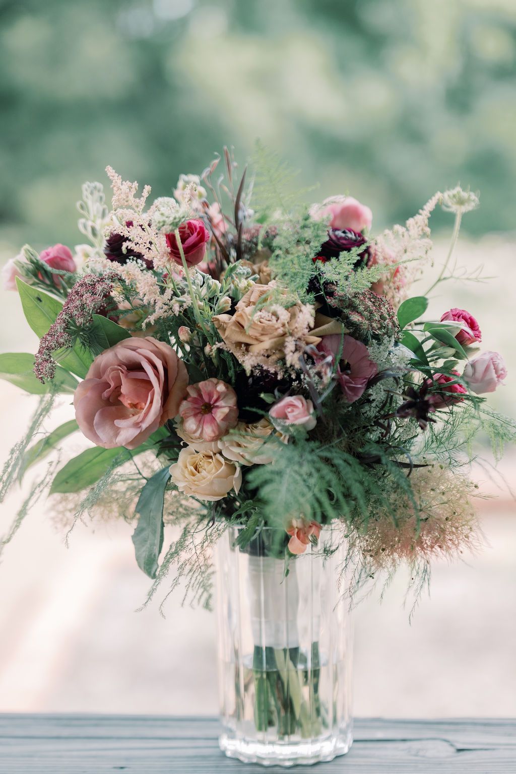 Elegant bridal bouquet of dusty pink roses, burgundy blooms, and wild greenery arranged in a clear glass vase.