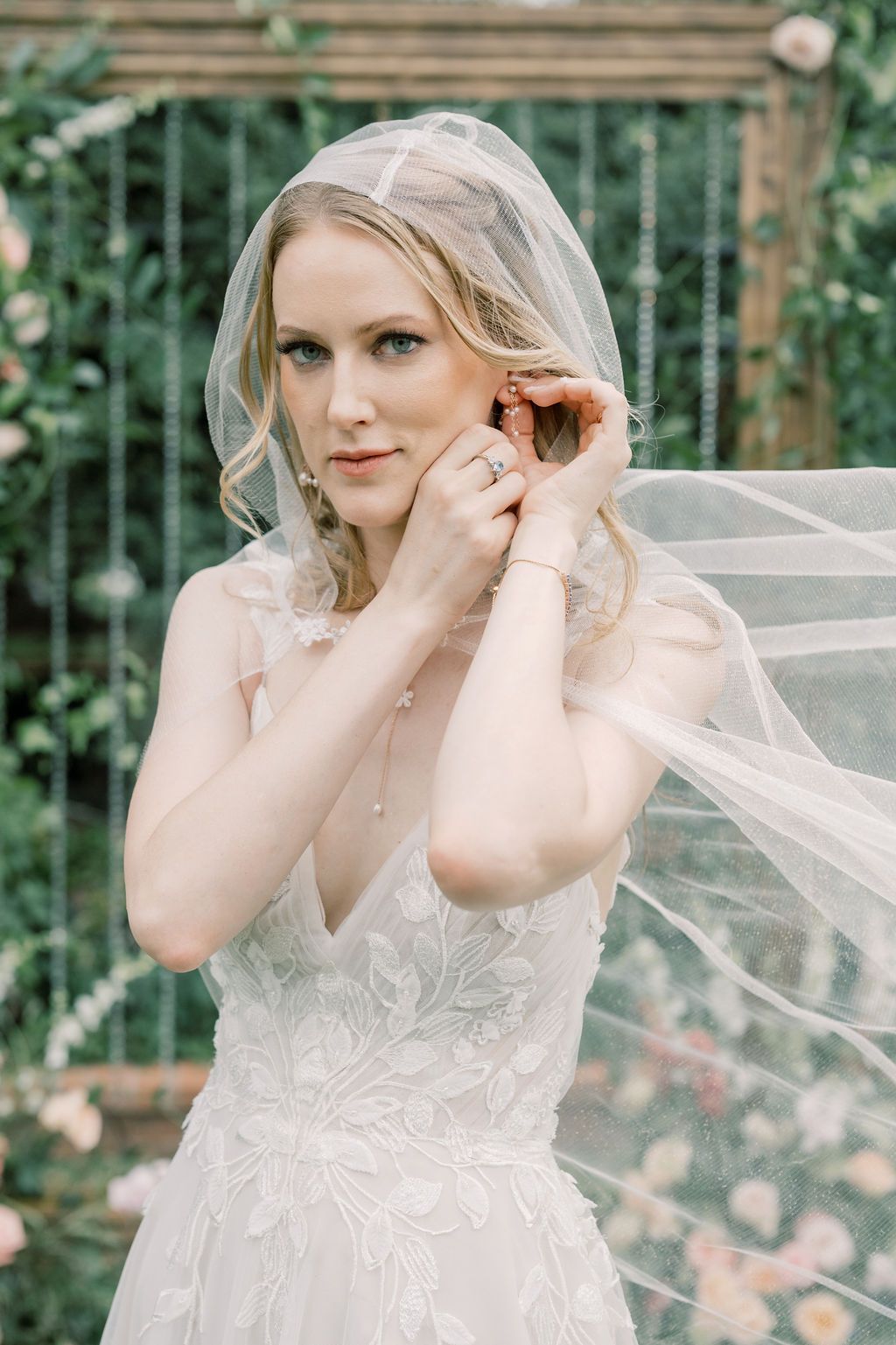 Blonde bride in lace wedding gown adjusting earring, wearing flowing veil, with floral garden backdrop.