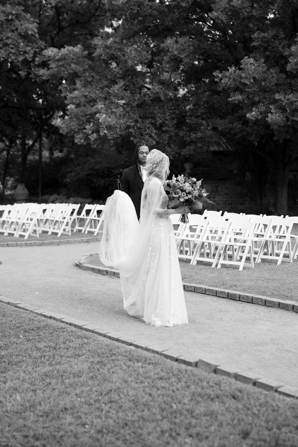Black and white photo of bride and groom kissing outdoors, white chairs lining aisle, bride holding bouquet.