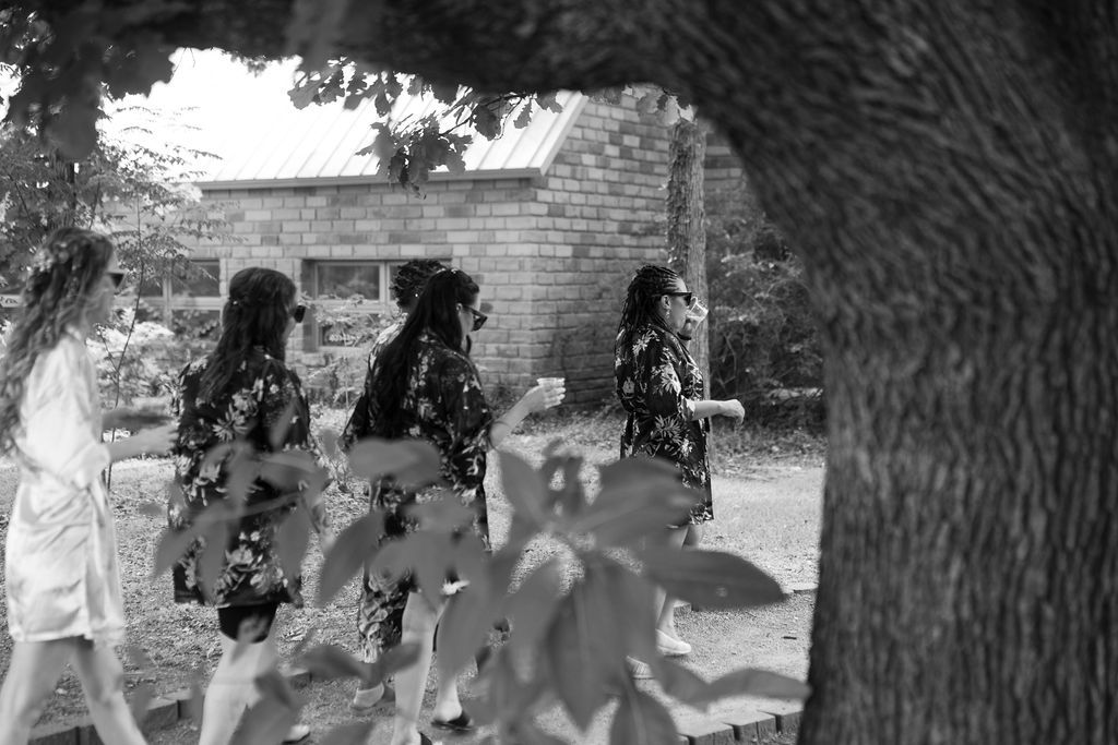Black and white photo of women in floral dresses walking outdoors near a brick building, framed by tree branches.