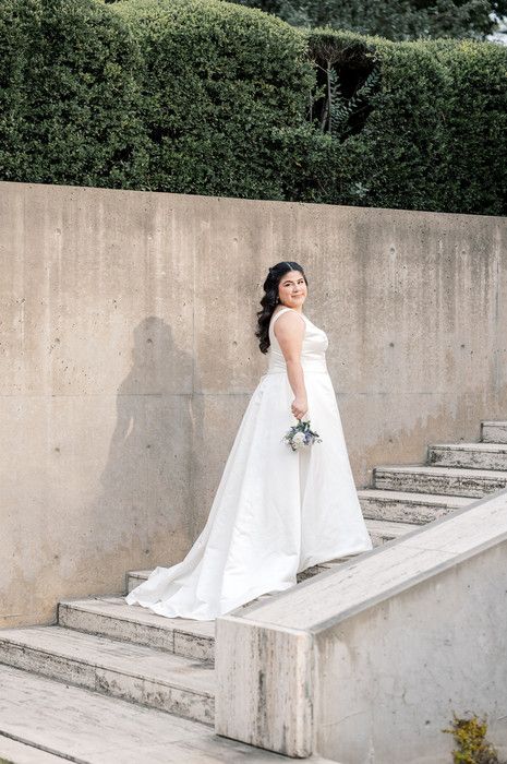Bride in white ball gown with train poses on marble steps with bouquet, concrete wall and hedges behind her.
