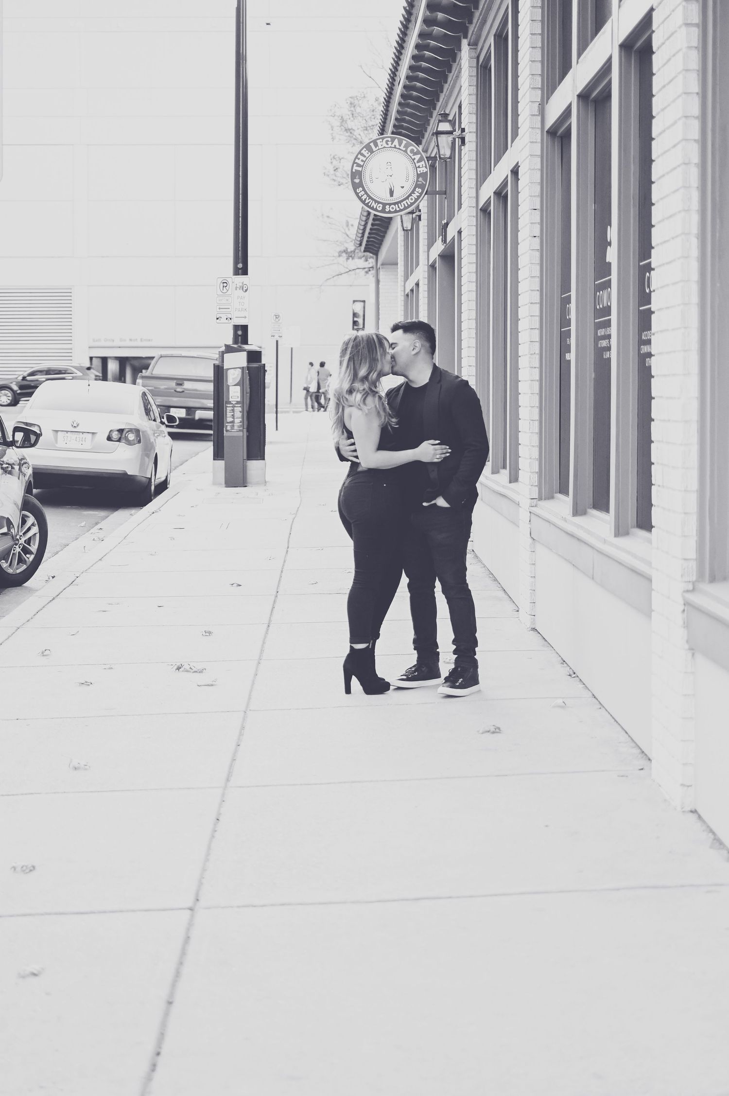 Black and white image of a couple sharing a kiss on an urban sidewalk near storefronts.