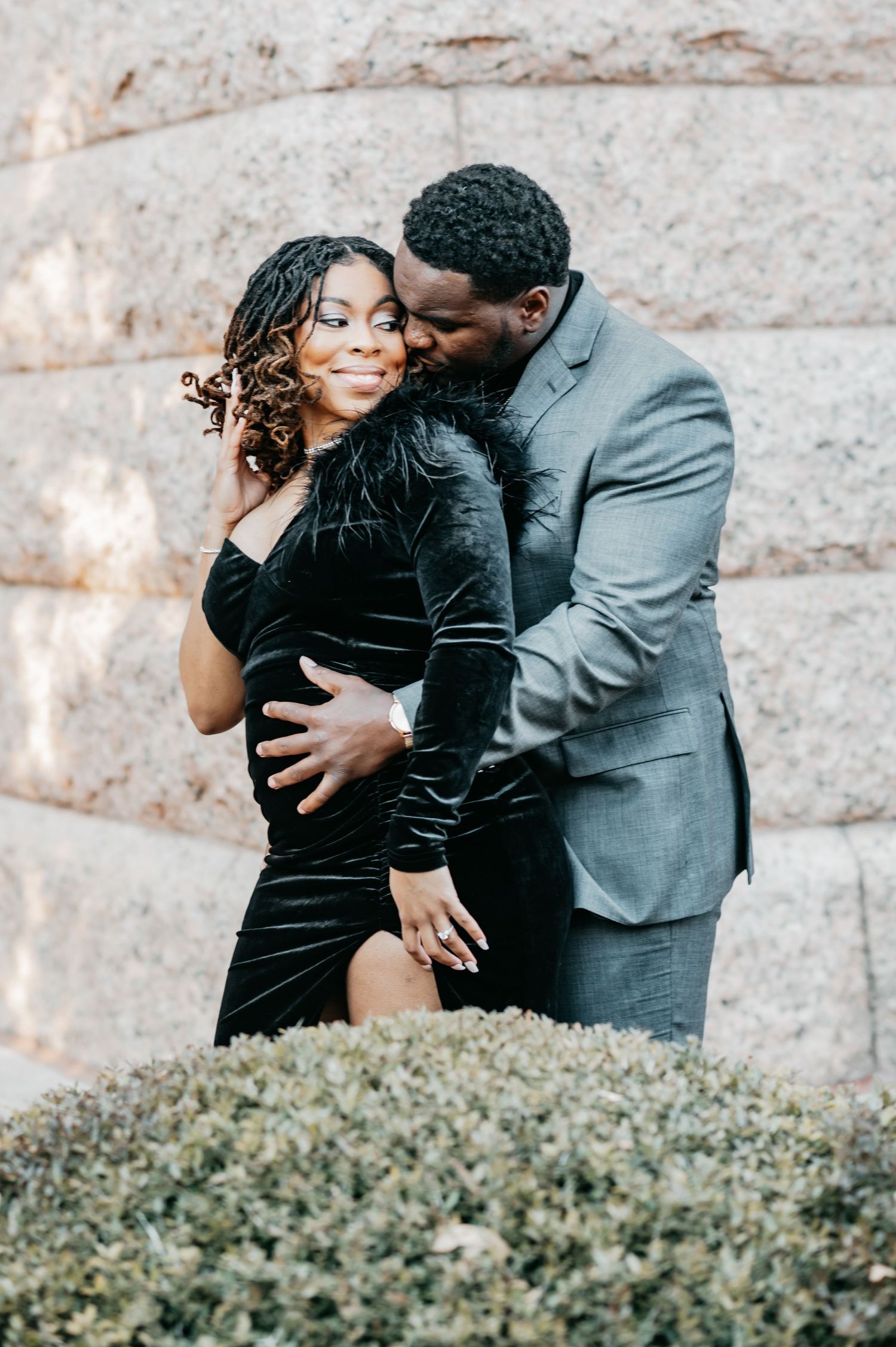 A couple shares a romantic embrace outdoors, woman in black velvet dress, man in gray suit, posing near stone wall.