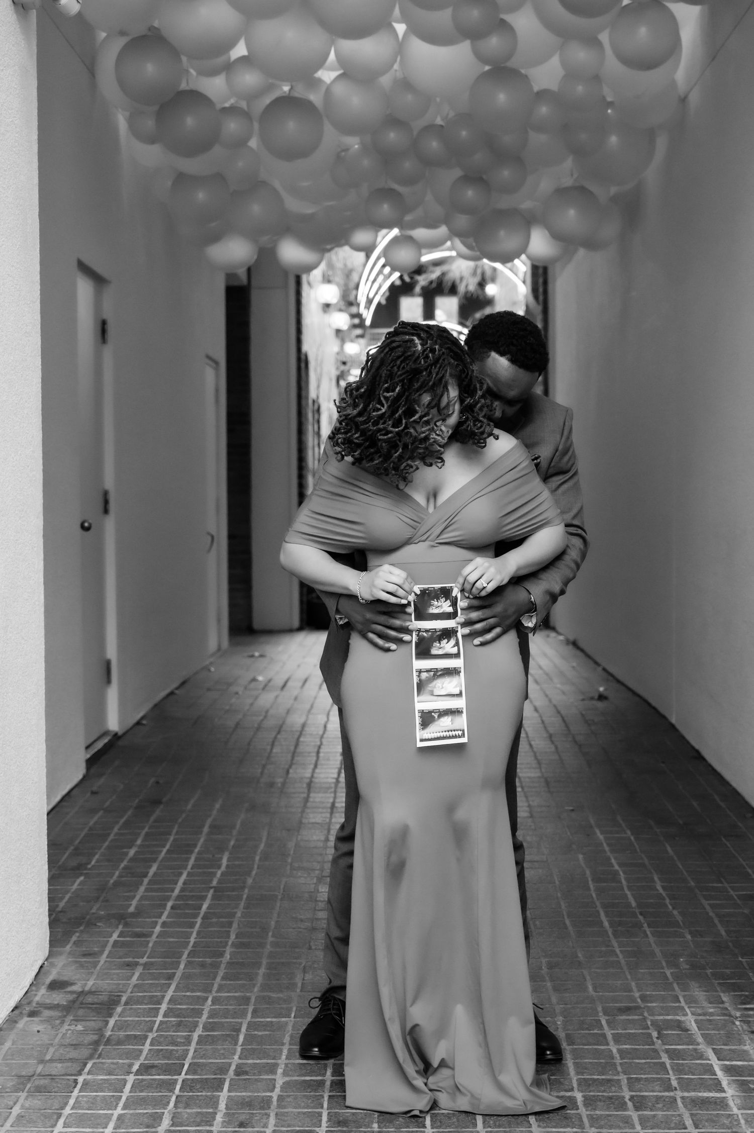 Black and white photo of a couple embracing in a balloon-filled hallway, holding ultrasound photos.