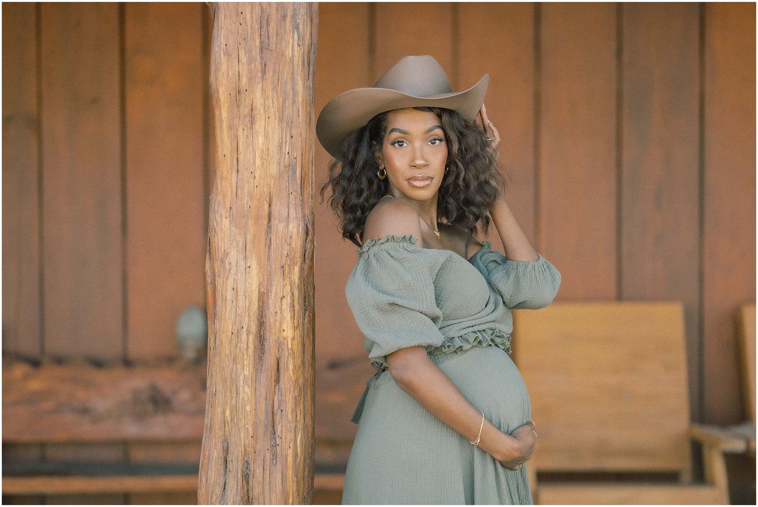 Maternity photoshoot featuring a sage green dress and brown hat against wooden walls.