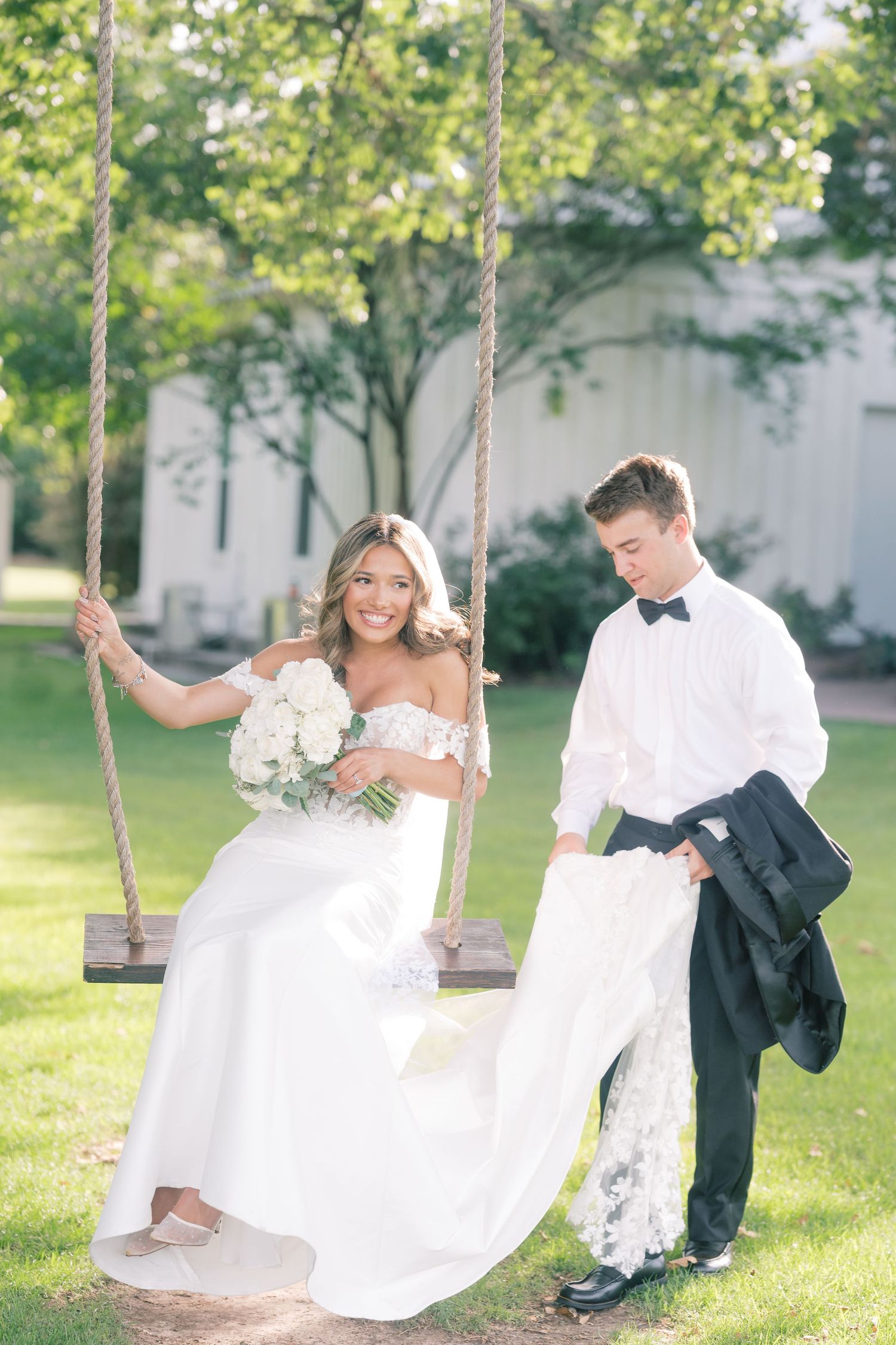 Smiling bride in off-shoulder gown sitting on rope swing holding bouquet while young man holds her train.
