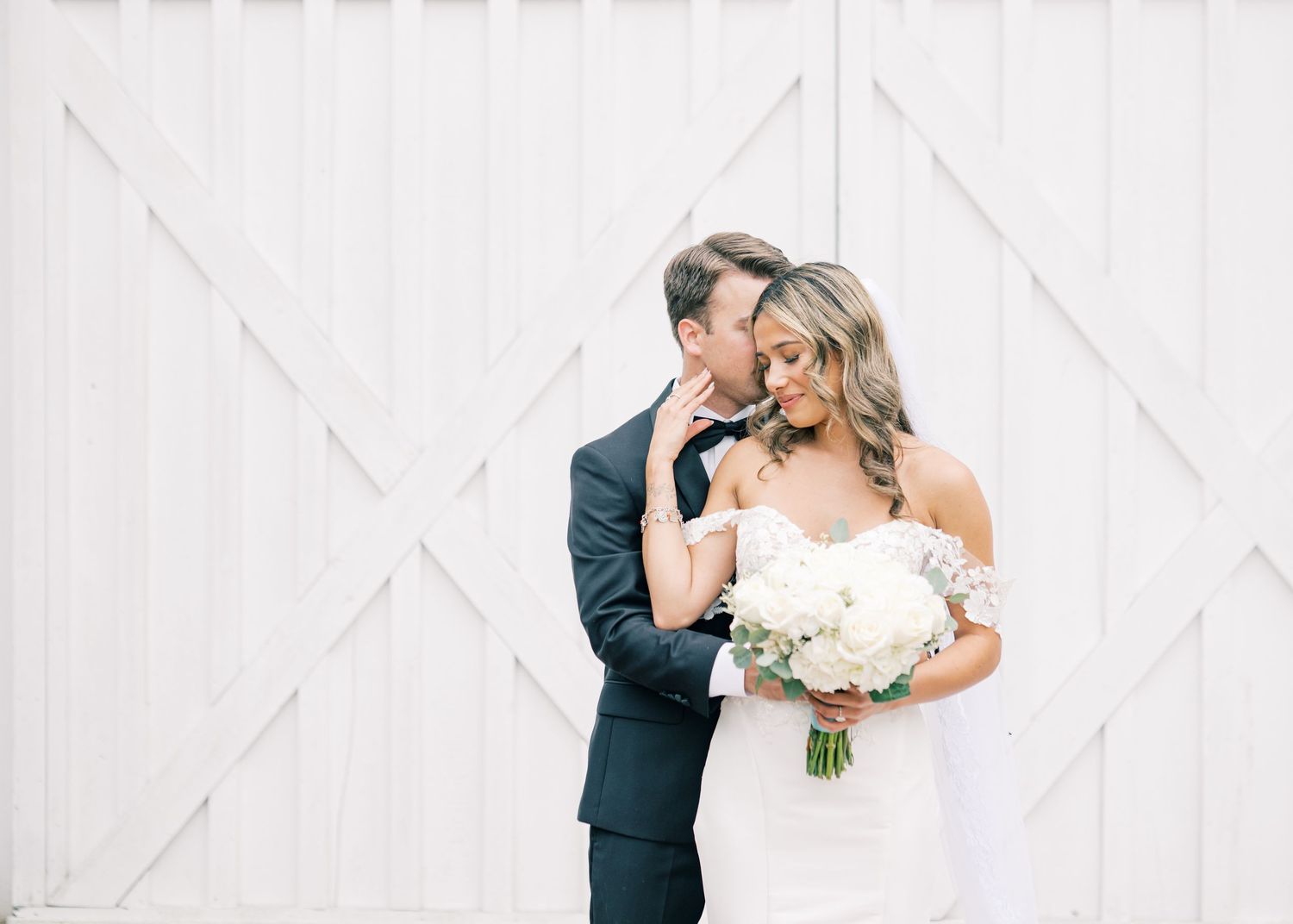Groom kisses bride on cheek as she smiles holding white bouquet in front of white barn doors.