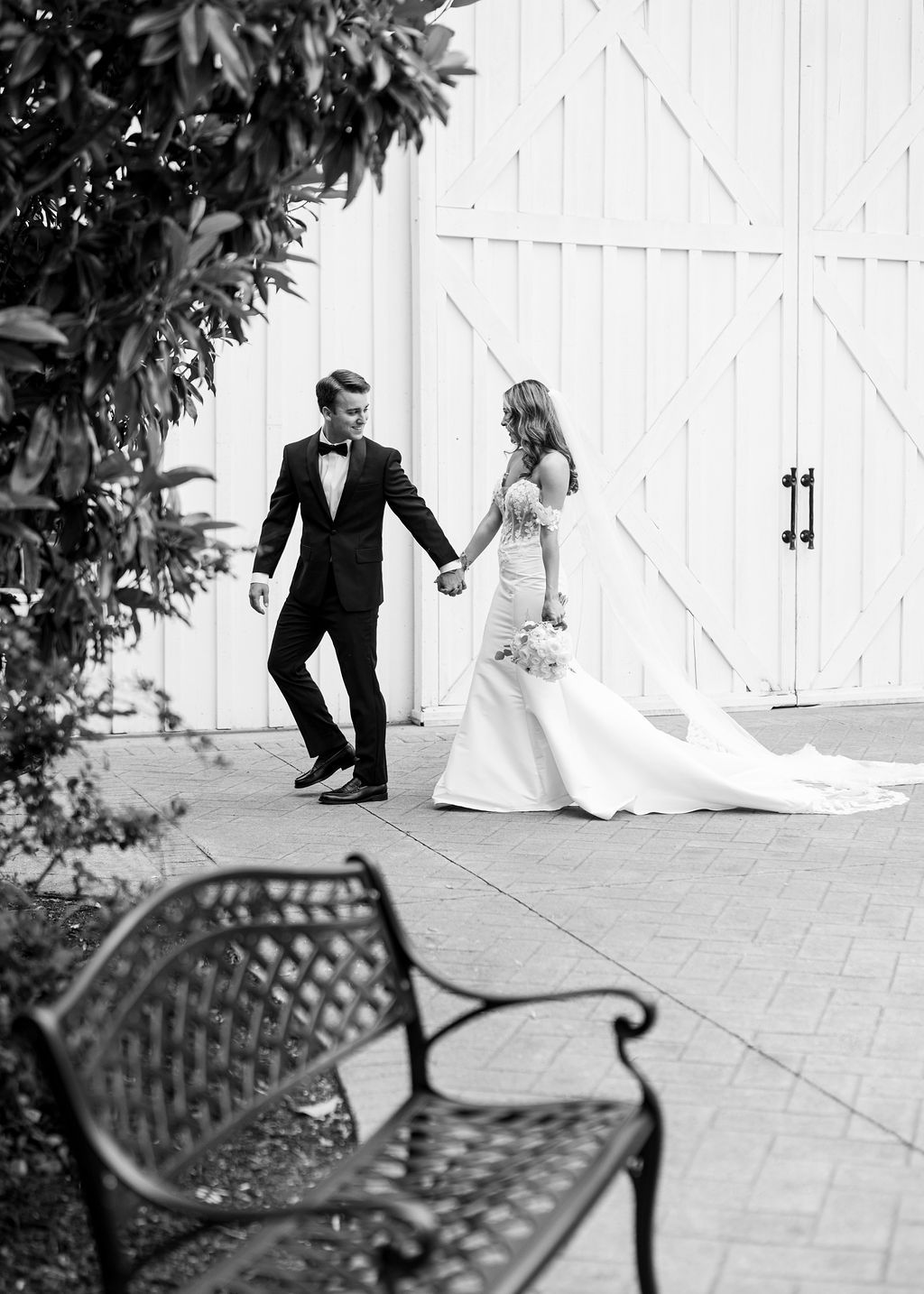 Black and white photo of bride and groom holding hands walking past white barn doors.