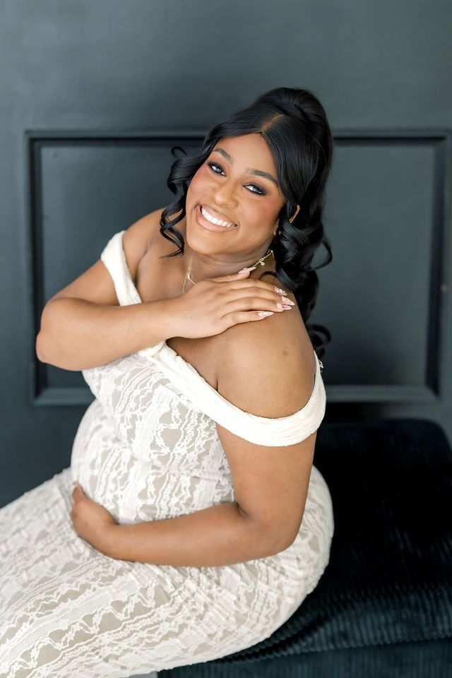 Smiling pregnant woman in white lace off-shoulder maternity gown poses against dark wall.