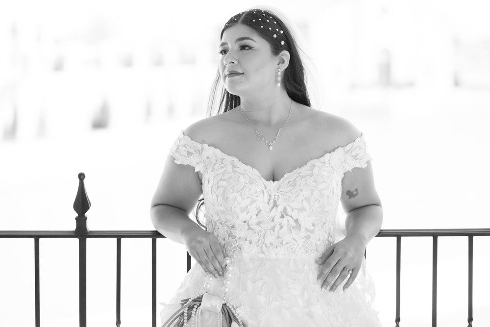 A bride in a white off-shoulder wedding gown poses elegantly against a stone wall backdrop.