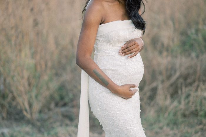 Side view of pregnant woman in fitted white dress highlighting her baby bump in nature setting.