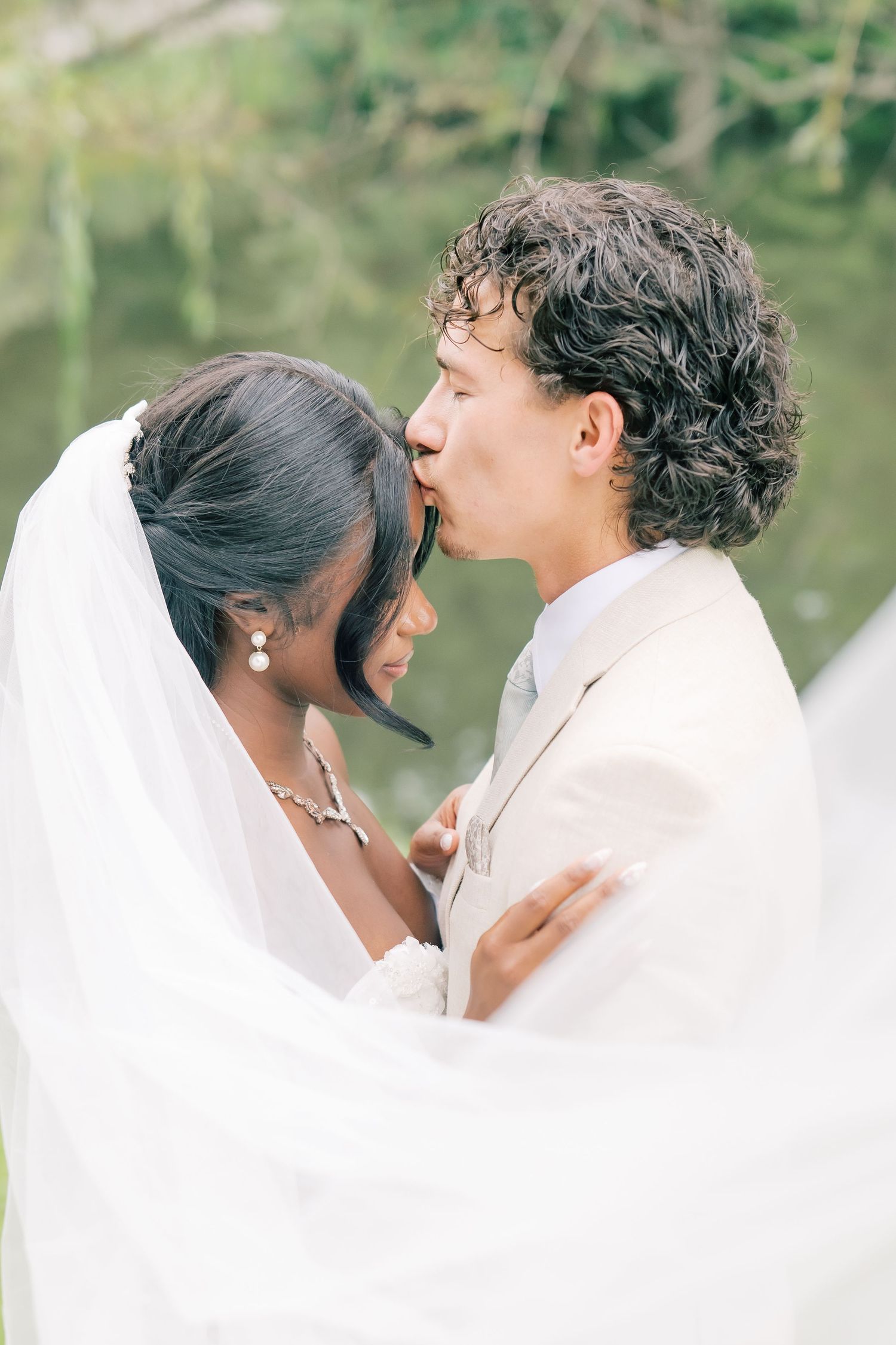 Bride and groom sharing tender forehead kiss under flowing white veil near a serene green pond backdrop.