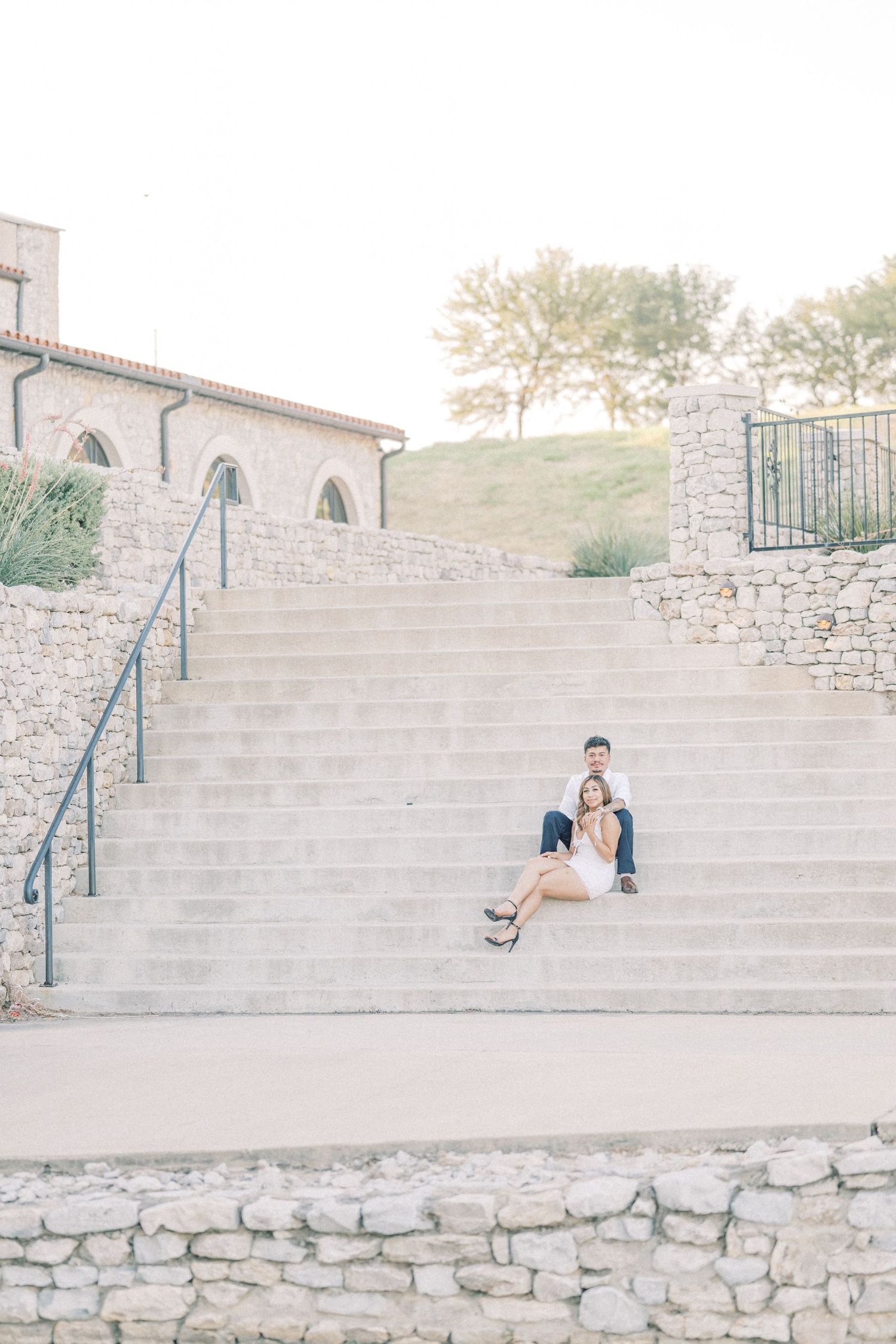 Engaged couple sitting together on wide stone steps outside a rustic venue at golden hour.