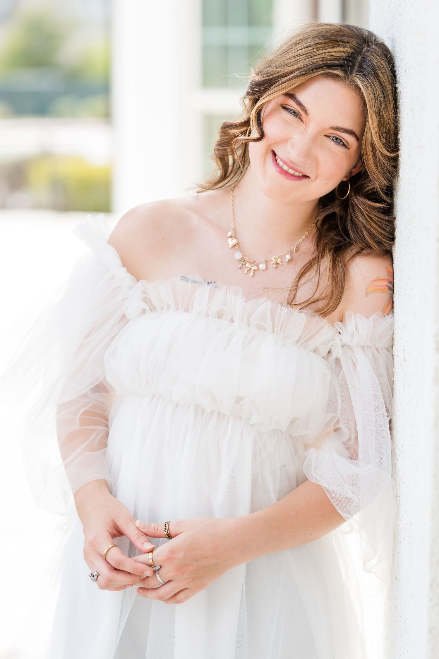Smiling woman in white off-shoulder tulle dress with pearl necklace leaning against a column.