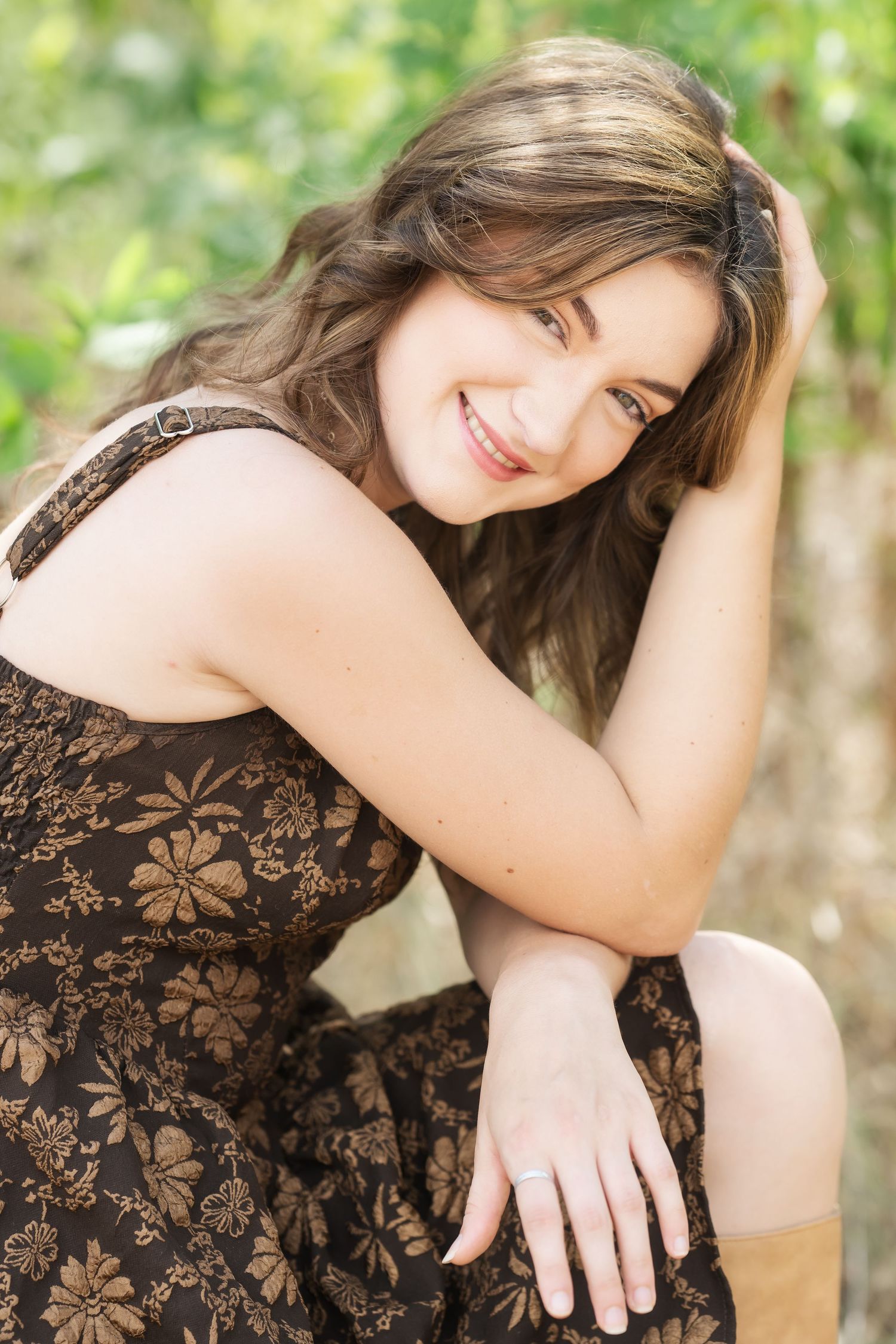 Smiling young woman in floral brown dress sitting outdoors with hand in hair against green foliage background.