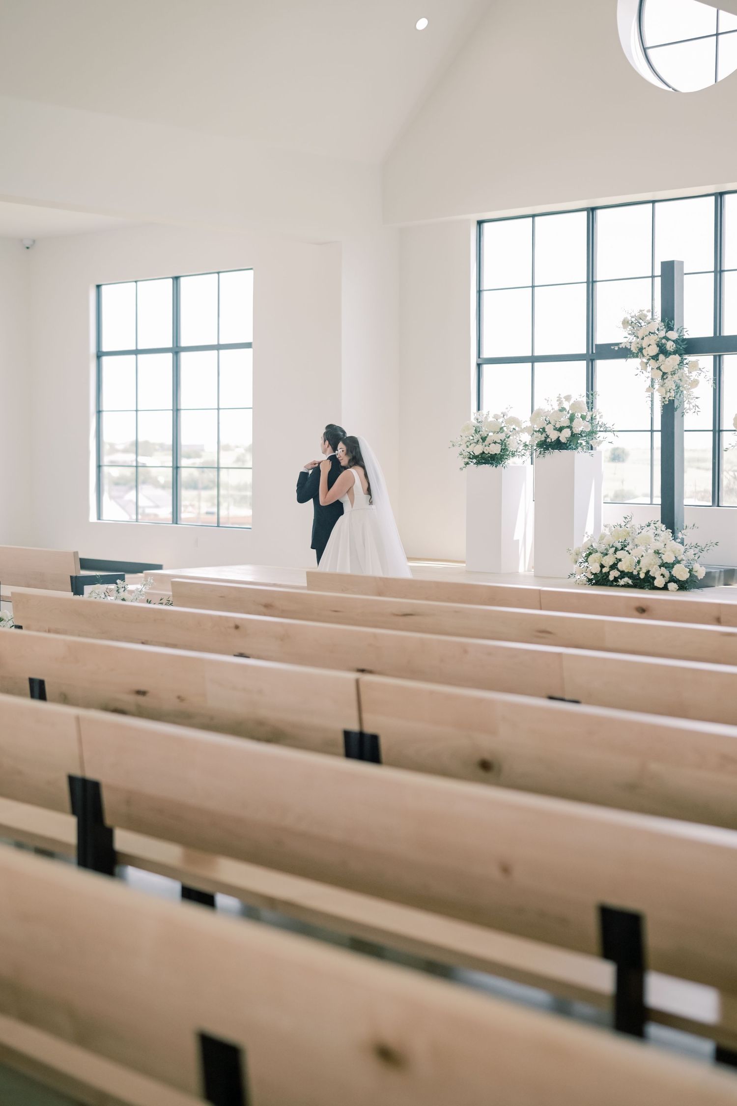 Bride and groom share an intimate embrace at the altar of a modern white chapel surrounded by elegant floral arrangements.