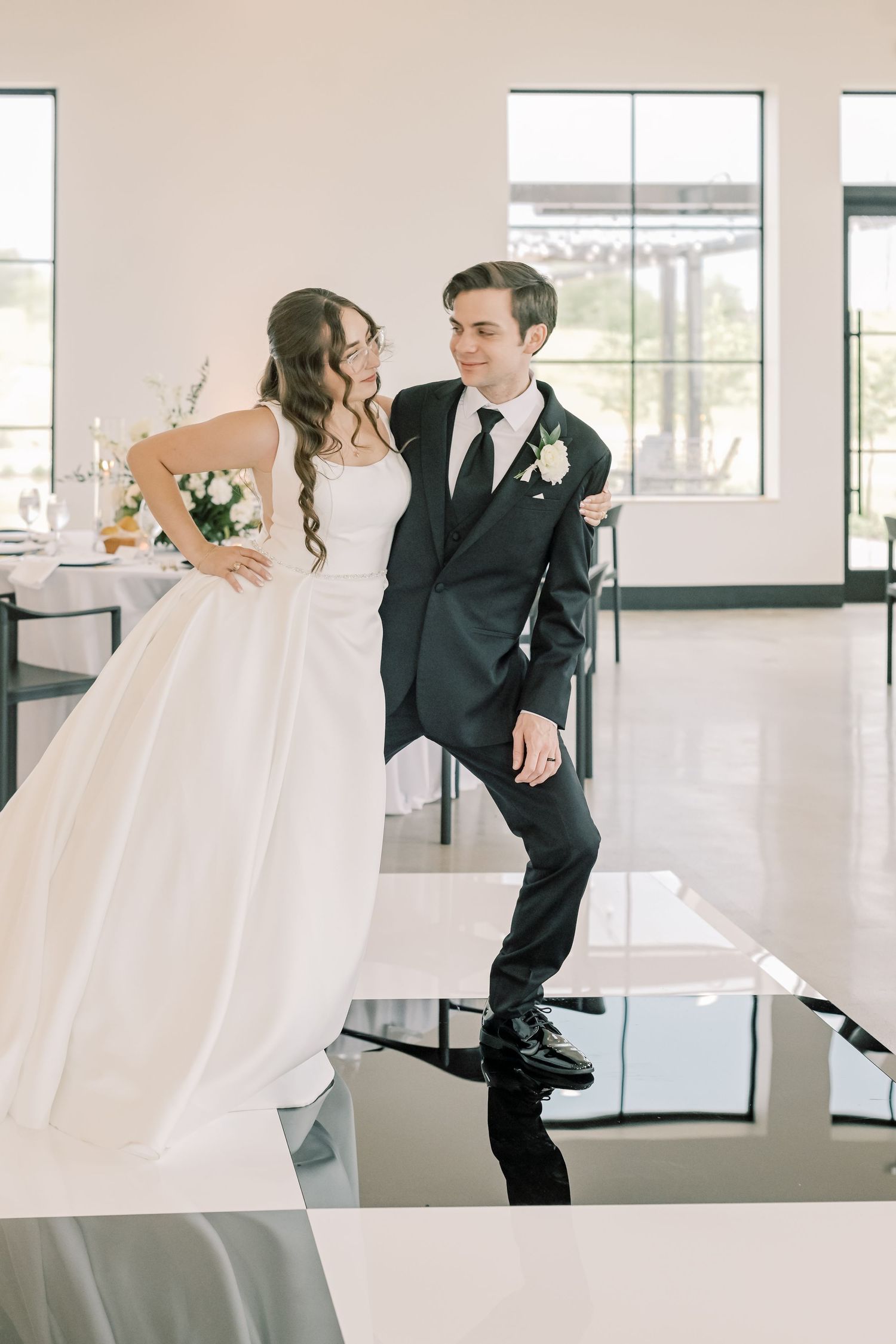 Bride in white ballgown and groom in black suit share a joyful moment in a bright wedding venue.