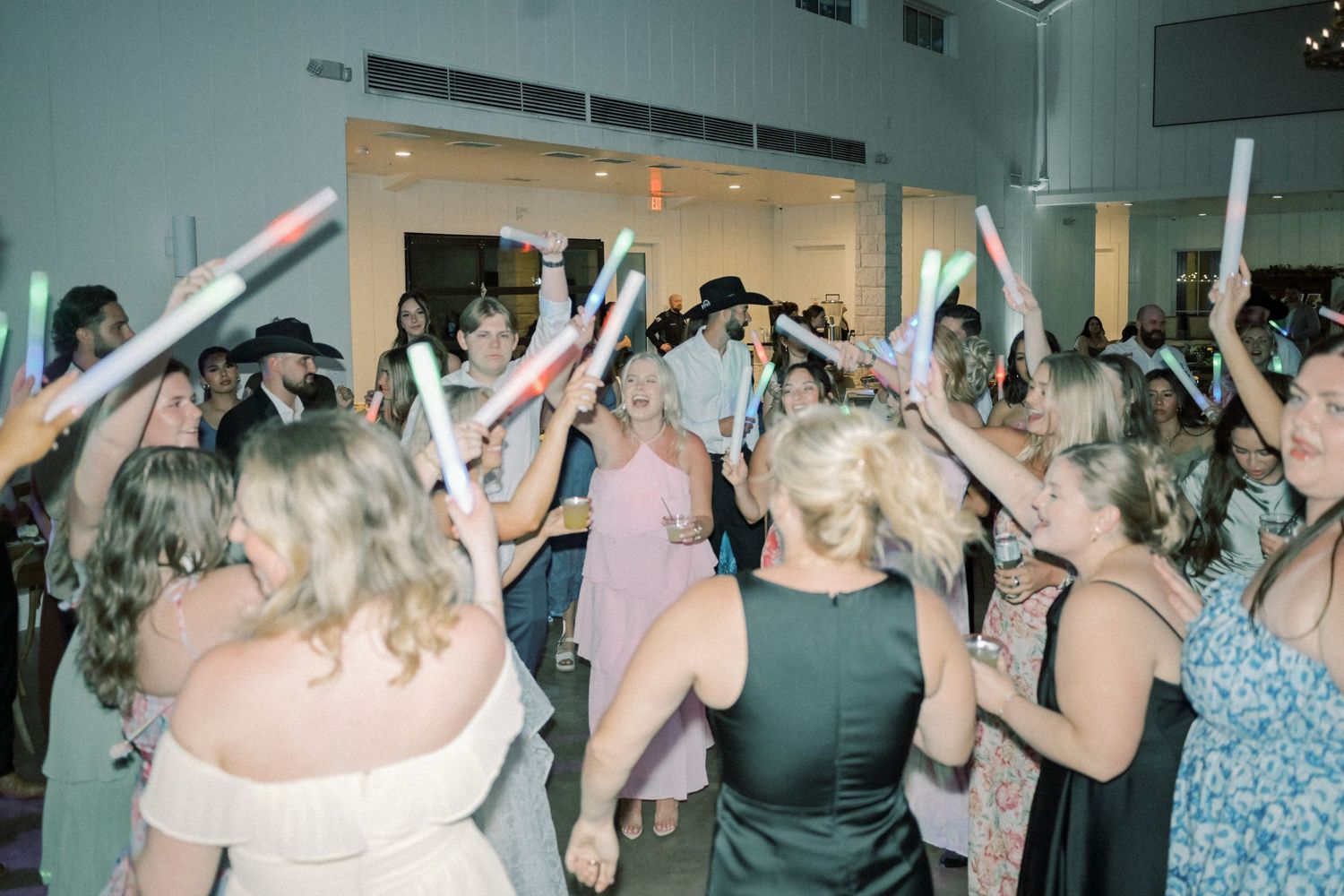 Excited wedding guests wave light-up foam sticks while dancing together in bright reception venue.
