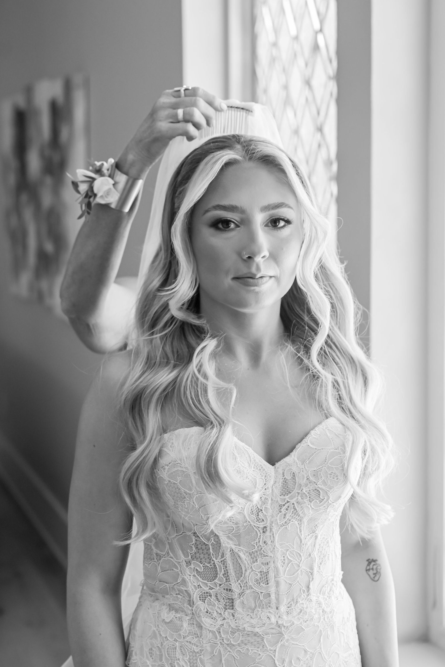 Black and white portrait of blonde bride in lace gown adjusting her veil by a window.