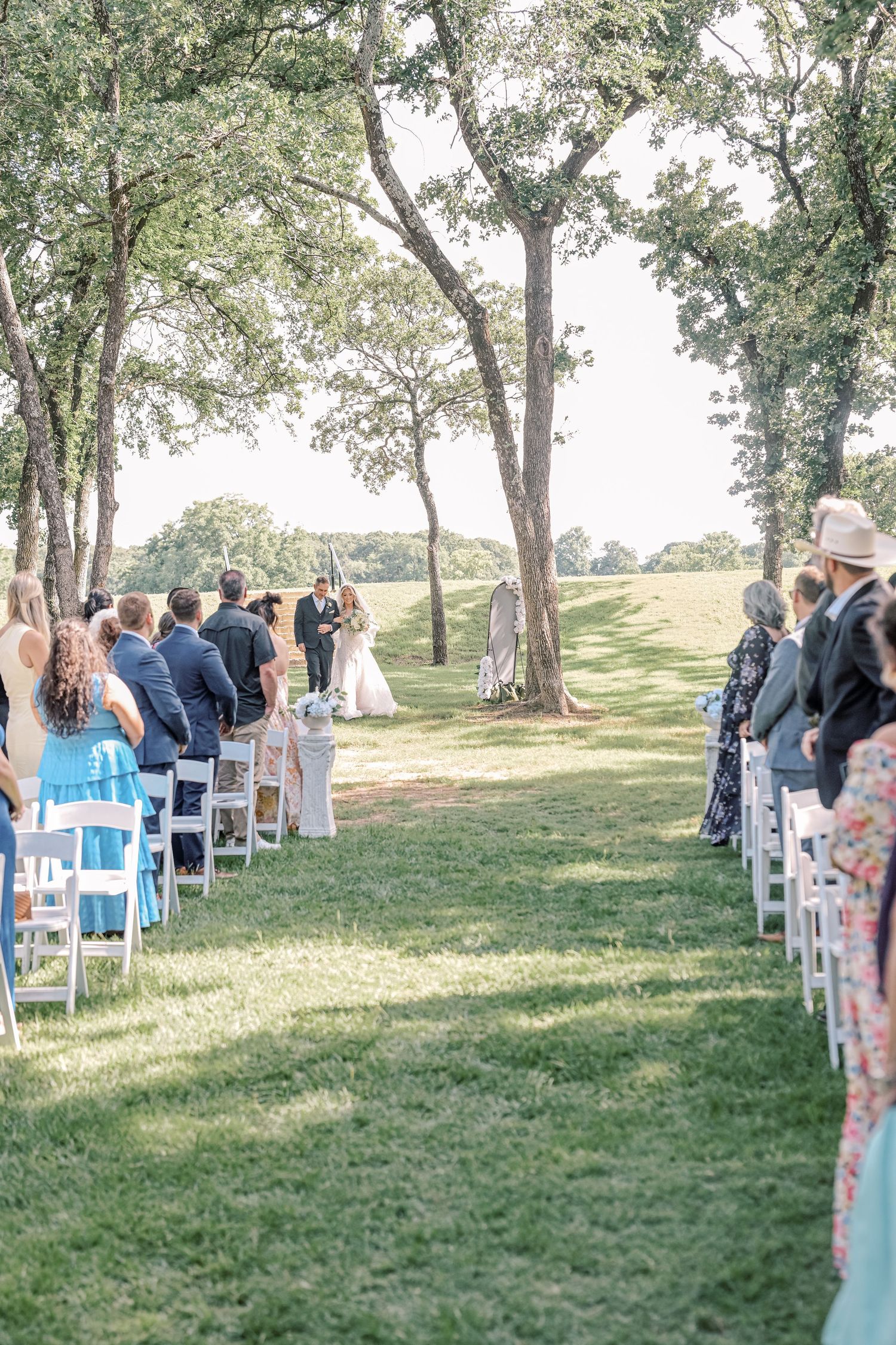Guests seated in white chairs watch outdoor wedding ceremony beneath towering oak trees.