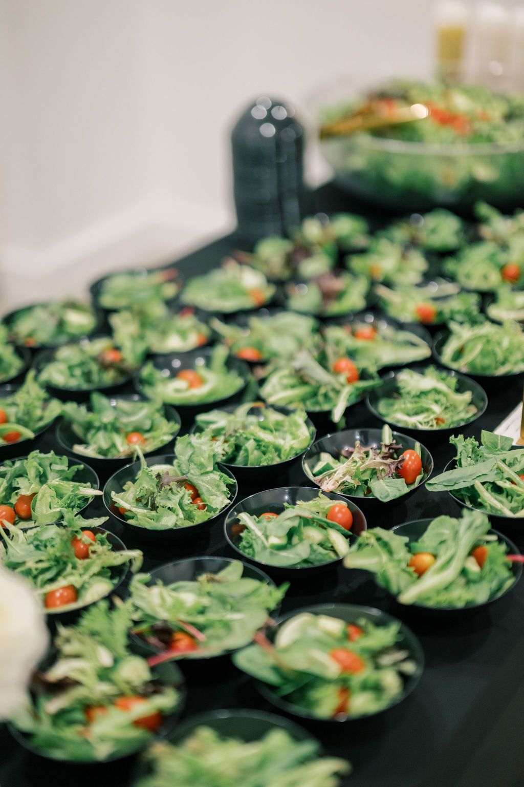 Multiple black bowls filled with fresh green salad and cherry tomatoes arranged on a dark table for catering.