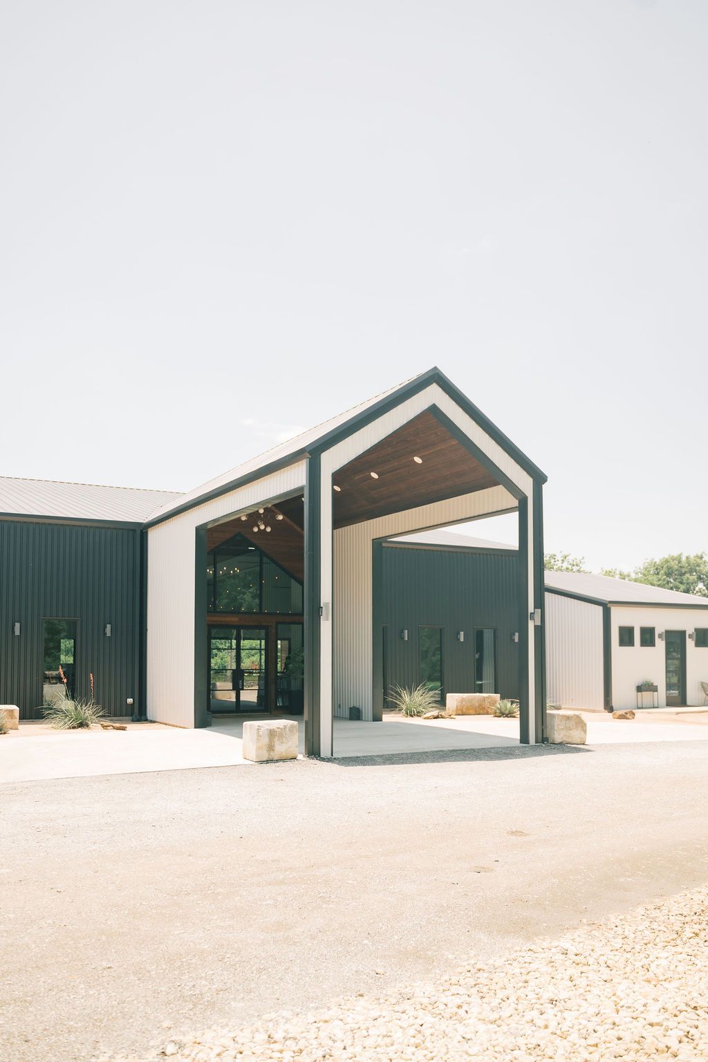 Modern barn-style wedding venue with dark green metal exterior, gabled entrance portico, and gravel driveway on a sunny day.