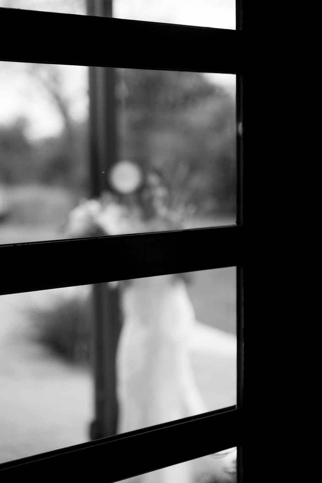 Artistic black and white photo of bride seen through window panes at wedding venue.